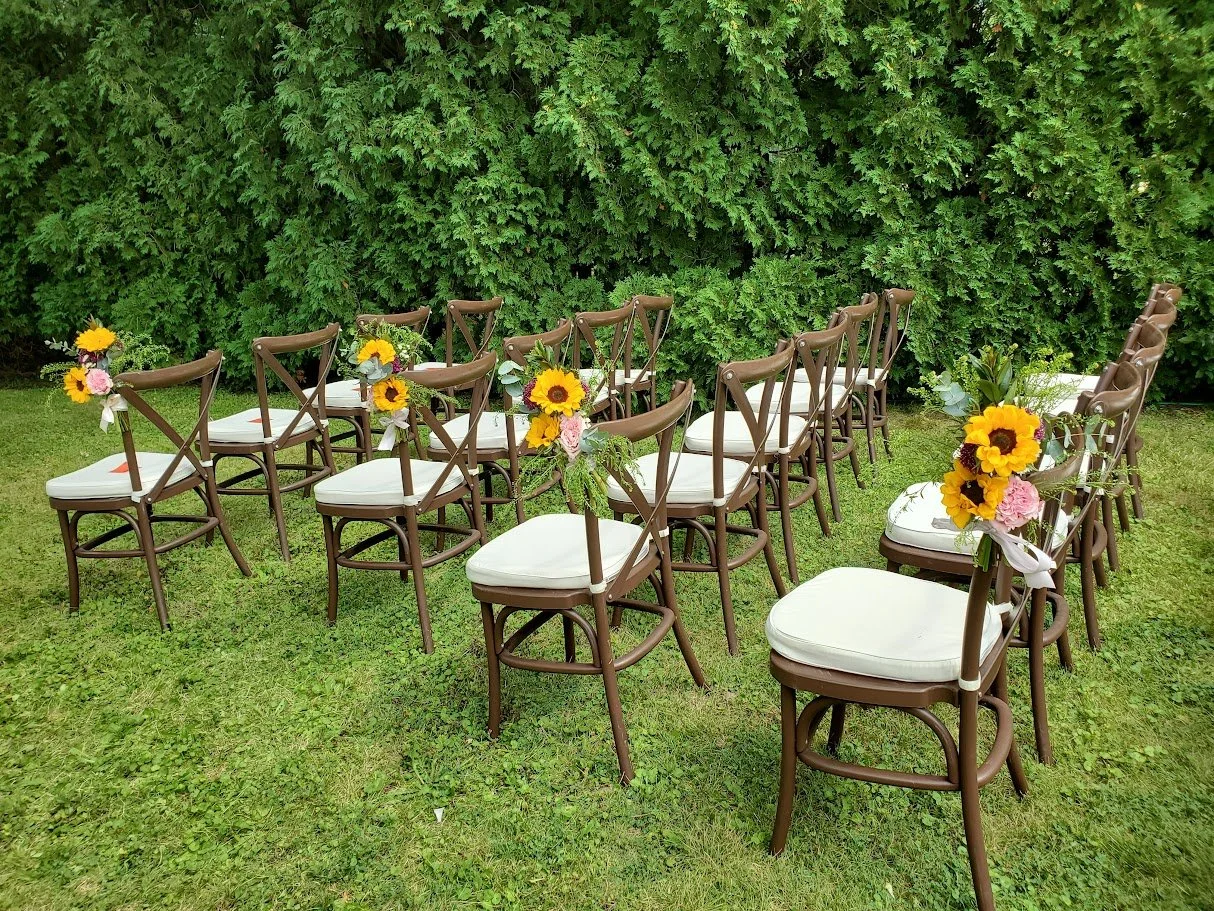 Outdoor wedding seating with brown chairs decorated with sunflower and pink flower bouquets, set on grass against a green leafy hedge.