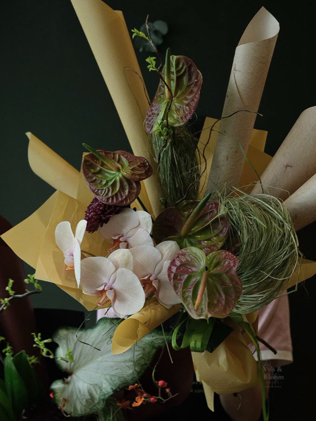 A bouquet of flowers including white orchids, patterned green and purple leaves, and green grasses, wrapped in yellow and brown paper.