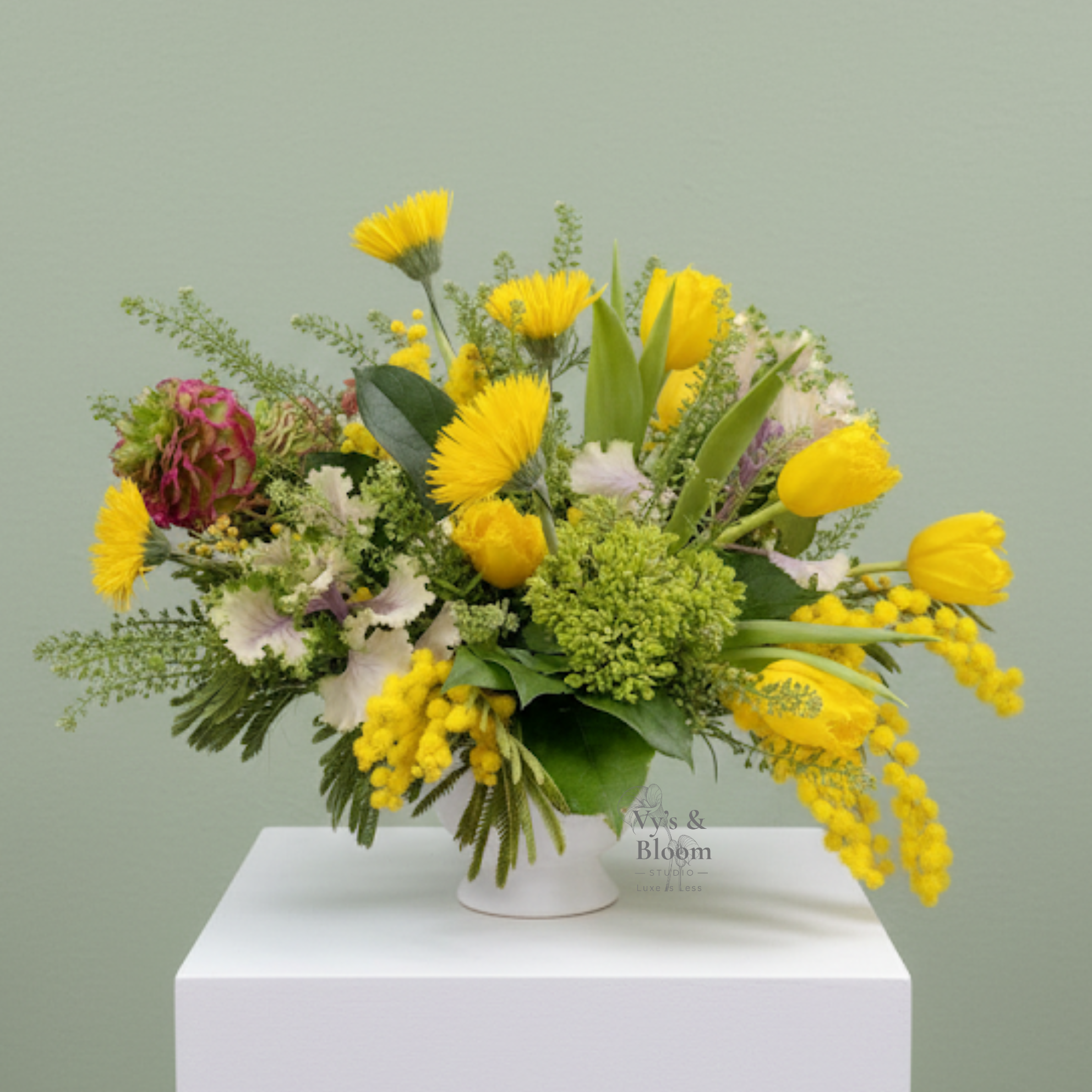 A floral arrangement featuring yellow tulips, yellow chrysanthemums, purple and white carnations, and green foliage on a white surface with a light green background.