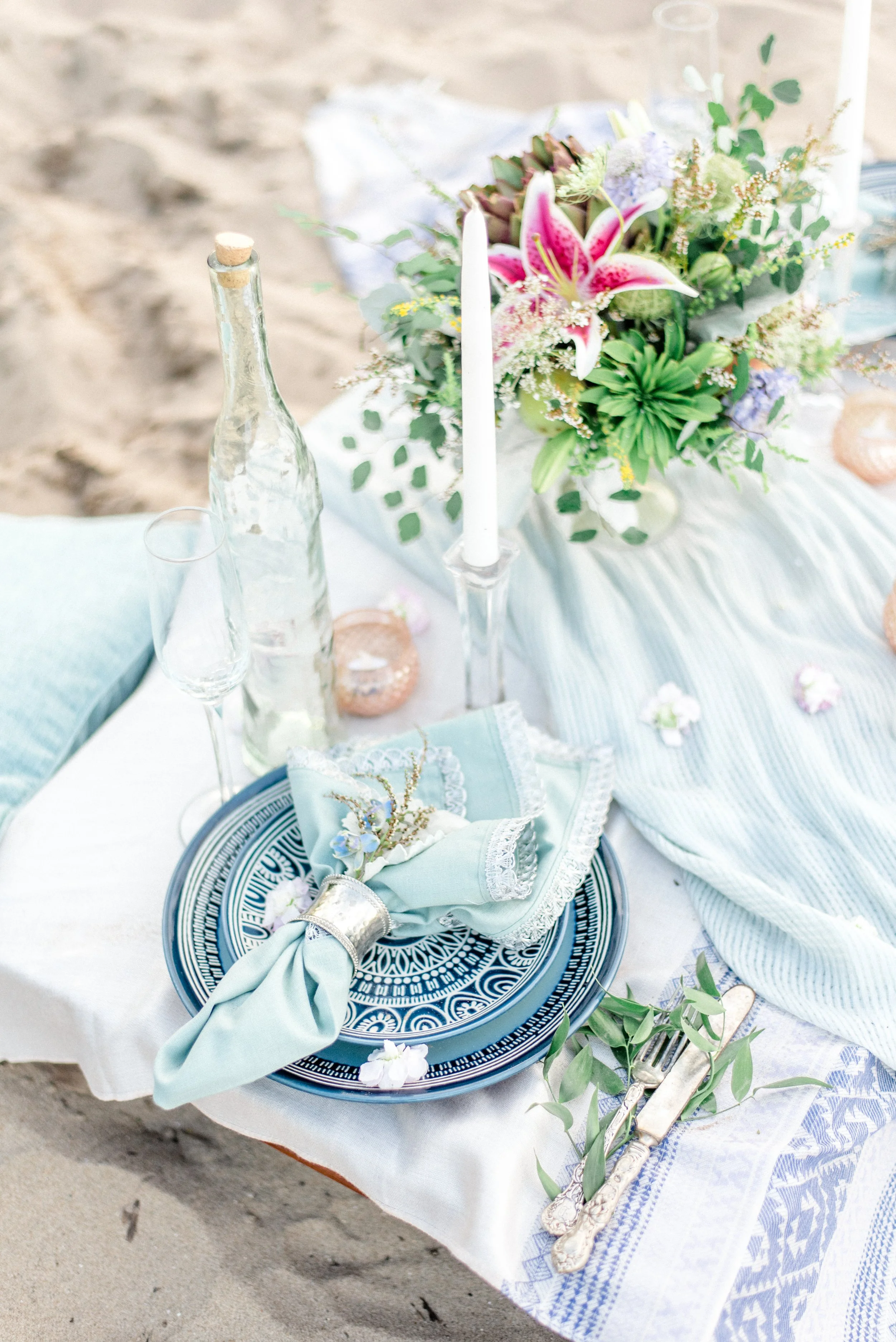 A beachside table setting with a floral centerpiece, candles, glass bottles, and tableware, arranged on a white cloth.