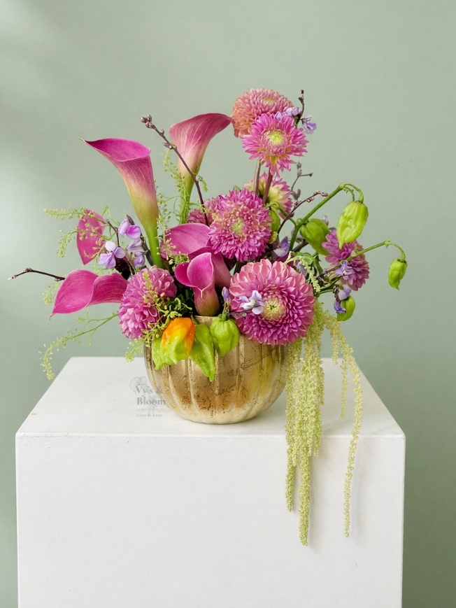Elegant floral arrangement with pink calla lilies, pink dahlias, and small purple and green flowers in a gold vase, placed on a white pedestal against a light green background.