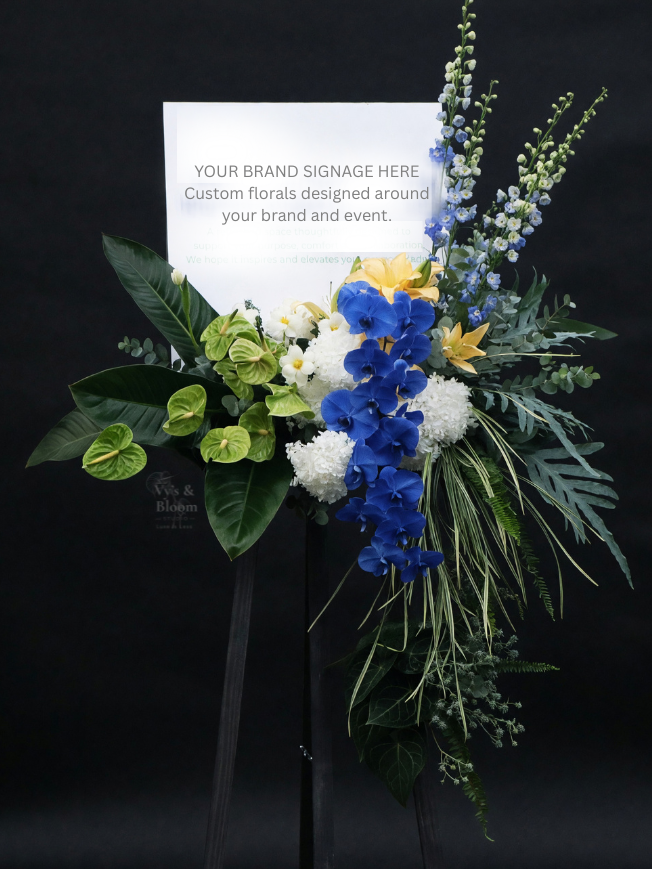 A floral arrangement with white, blue, yellow, and green flowers on a stand, with a sign that reads 'Your Brand Signage Here' for a brand or event display.