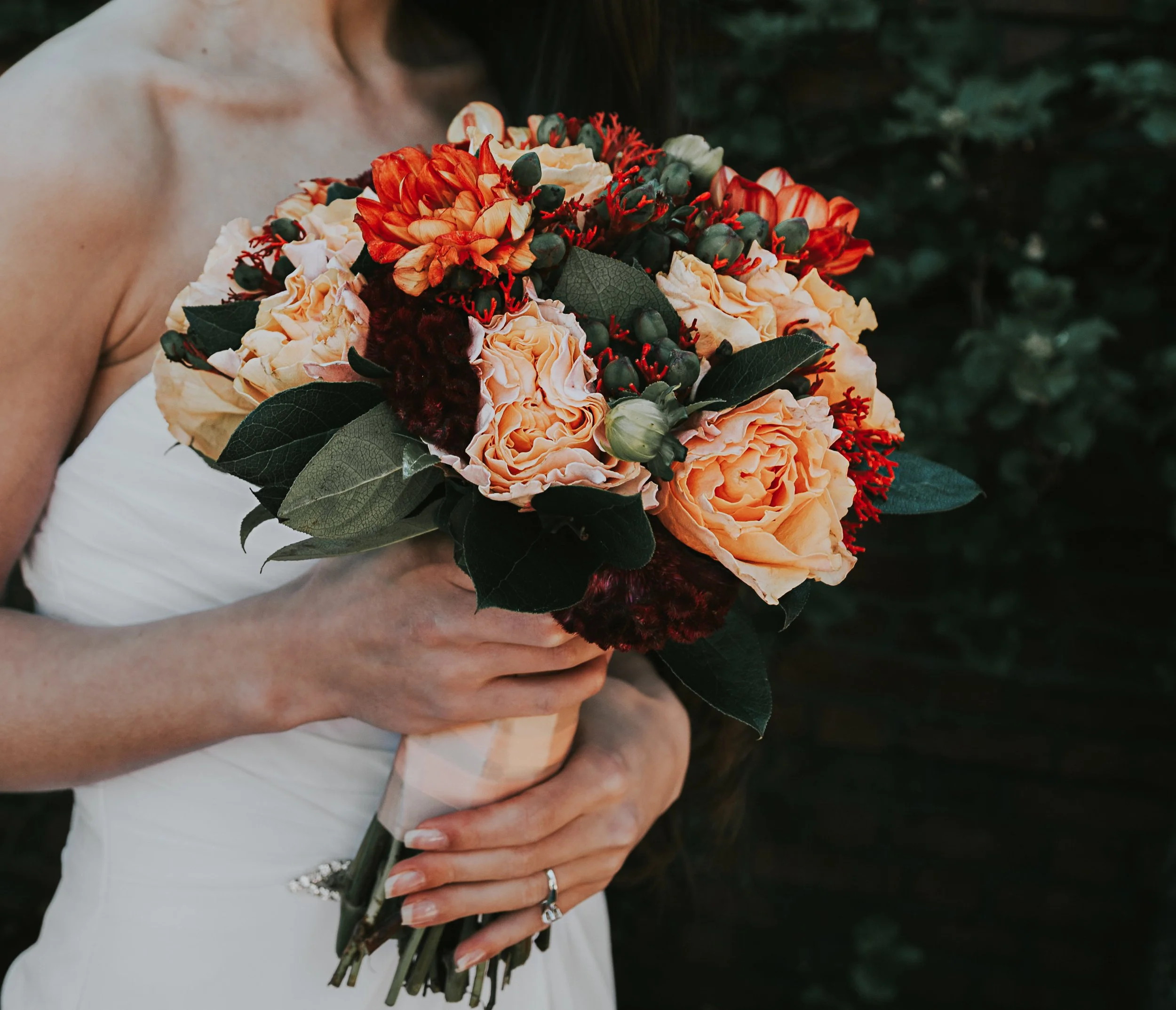 A person holding a bouquet of peach, red, and white flowers with green leaves outdoors.