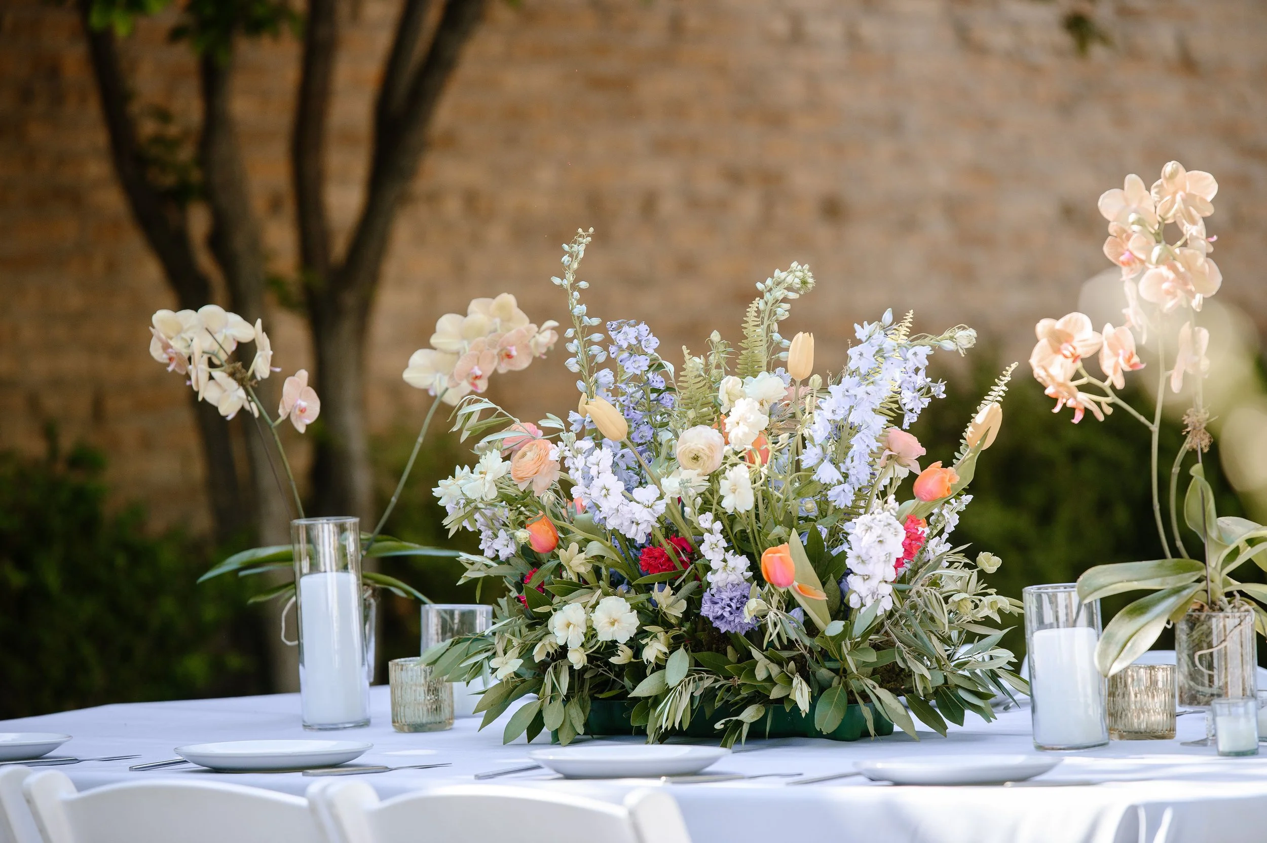 A large floral centerpiece with white, pink, purple, and peach flowers on a table set for an outdoor event.