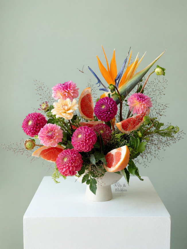 A floral arrangement with pink and purple dahlias, orange and purple bird of paradise flowers, and slices of grapefruit in a white vase on a white pedestal against a light-colored background.