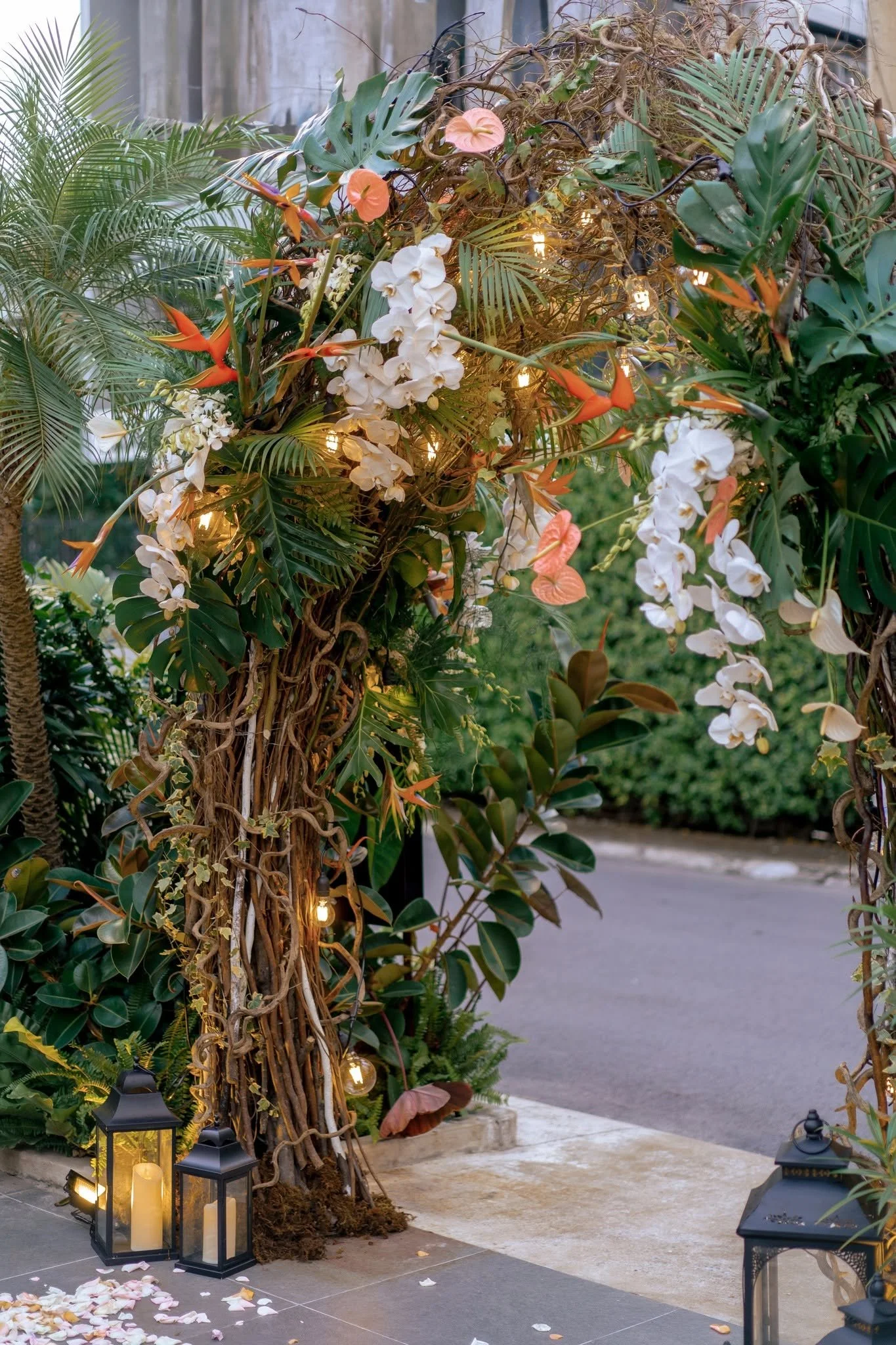 Decorative outdoor wedding or ceremony setup with lush greenery, hanging flowers, lanterns, and scattered flower petals.