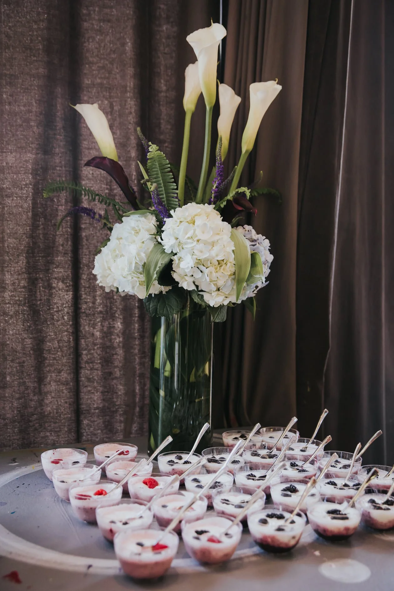 A tall glass vase with white calla lilies, hydrangeas, and purple accents on a table. In front, there are small cups of dessert with red woven decorations and spoons.