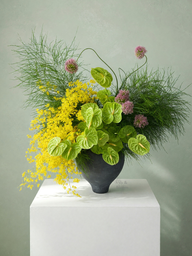 A black vase with an arrangement of yellow, pink, and green flowers and foliage, displayed on a white pedestal.