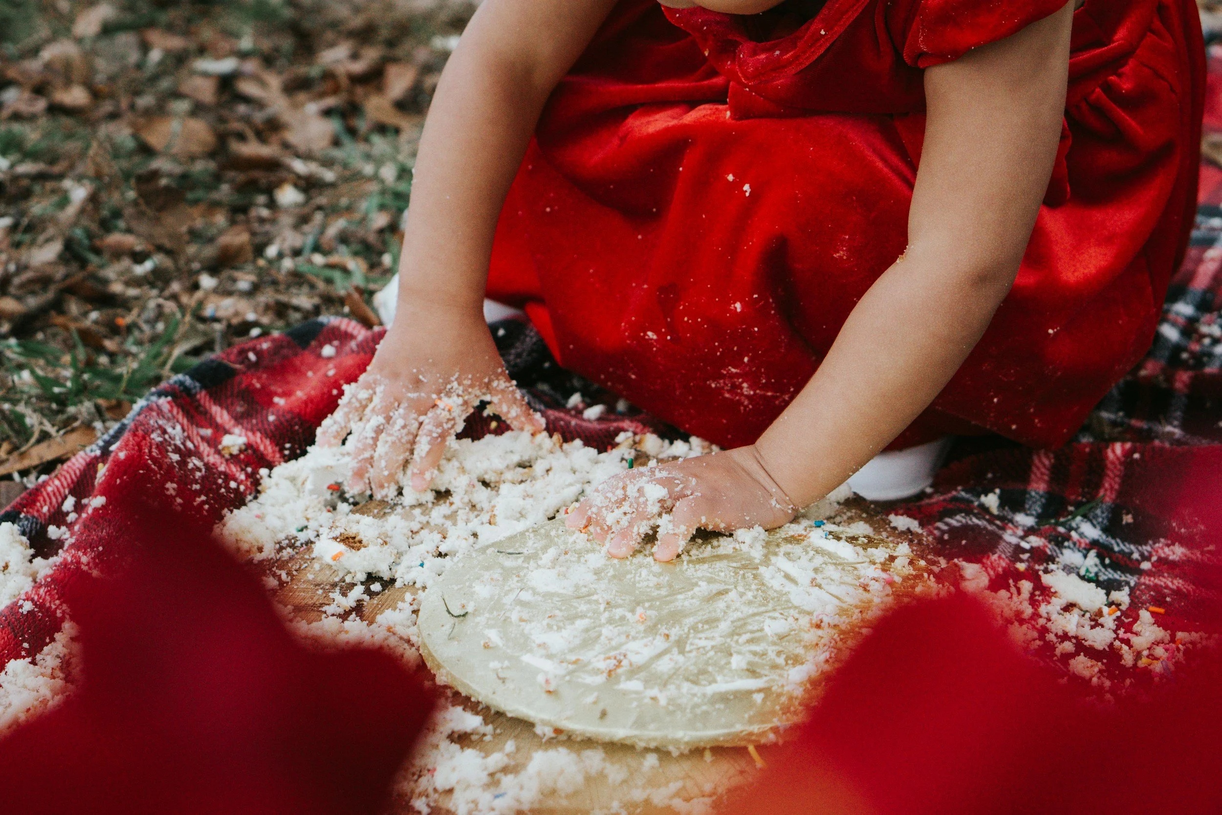 A child wearing a red dress and red shoes sitting outdoors on the ground, playing with a dough-like substance and making snow angels on a red and black checkered blanket.