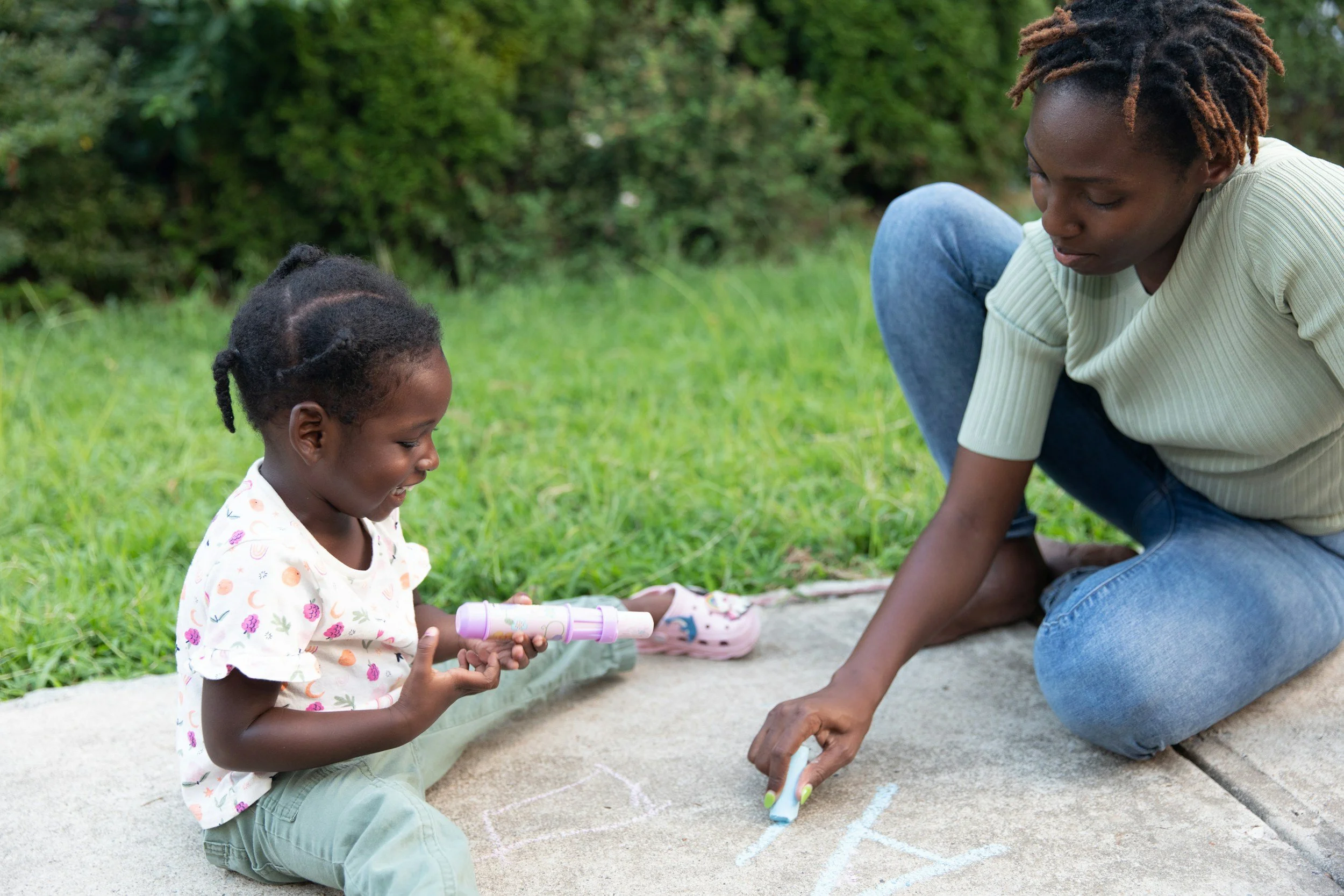 A woman and a young girl drawing with chalk on the sidewalk outdoors, with grass and bushes in the background.