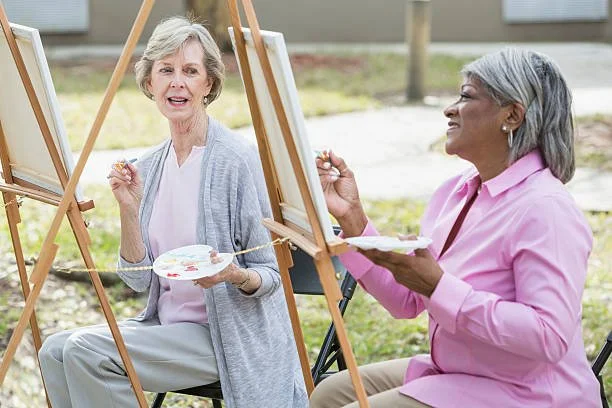 Two elderly women painting on outdoor easels and smiling at each other.