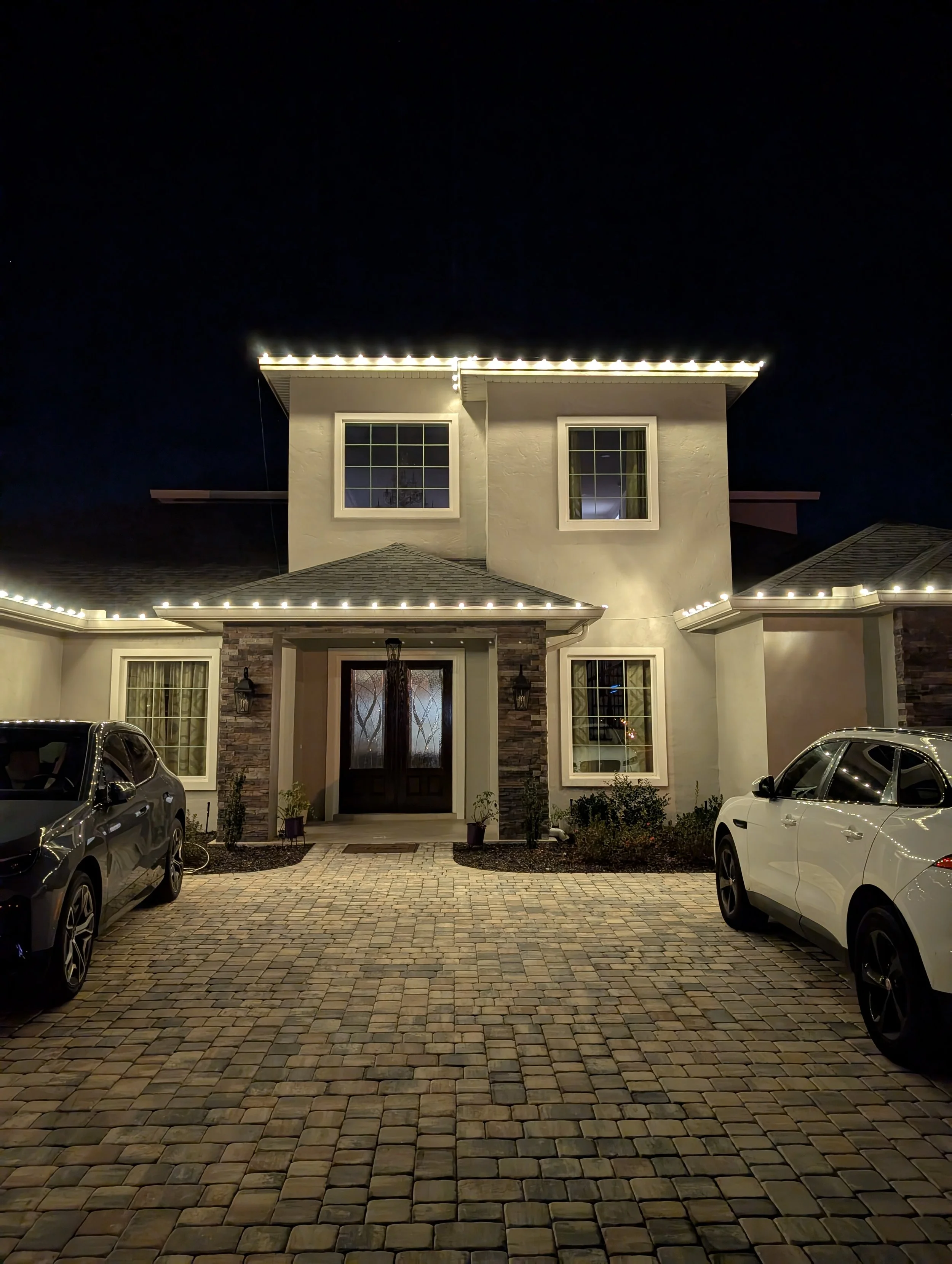 A modern two-story house at night with exterior lights outlining the roof and windows. The house has a stone and stucco facade with a driveway and parked cars in front. Orlando Christmas Lights, Orange County Holiday Lighting, Best Christmas Lights