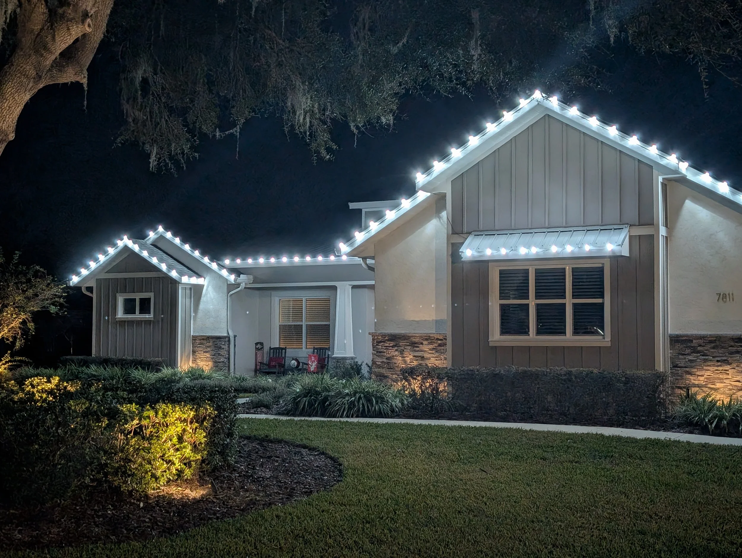 A farmhouse-style home at night with bright white string lights along the roofline and landscape uplighting on the front bushes. Orlando Christmas Lights, Winter Park Holiday Lighting, Windermere Christmas Light Installation Near Me