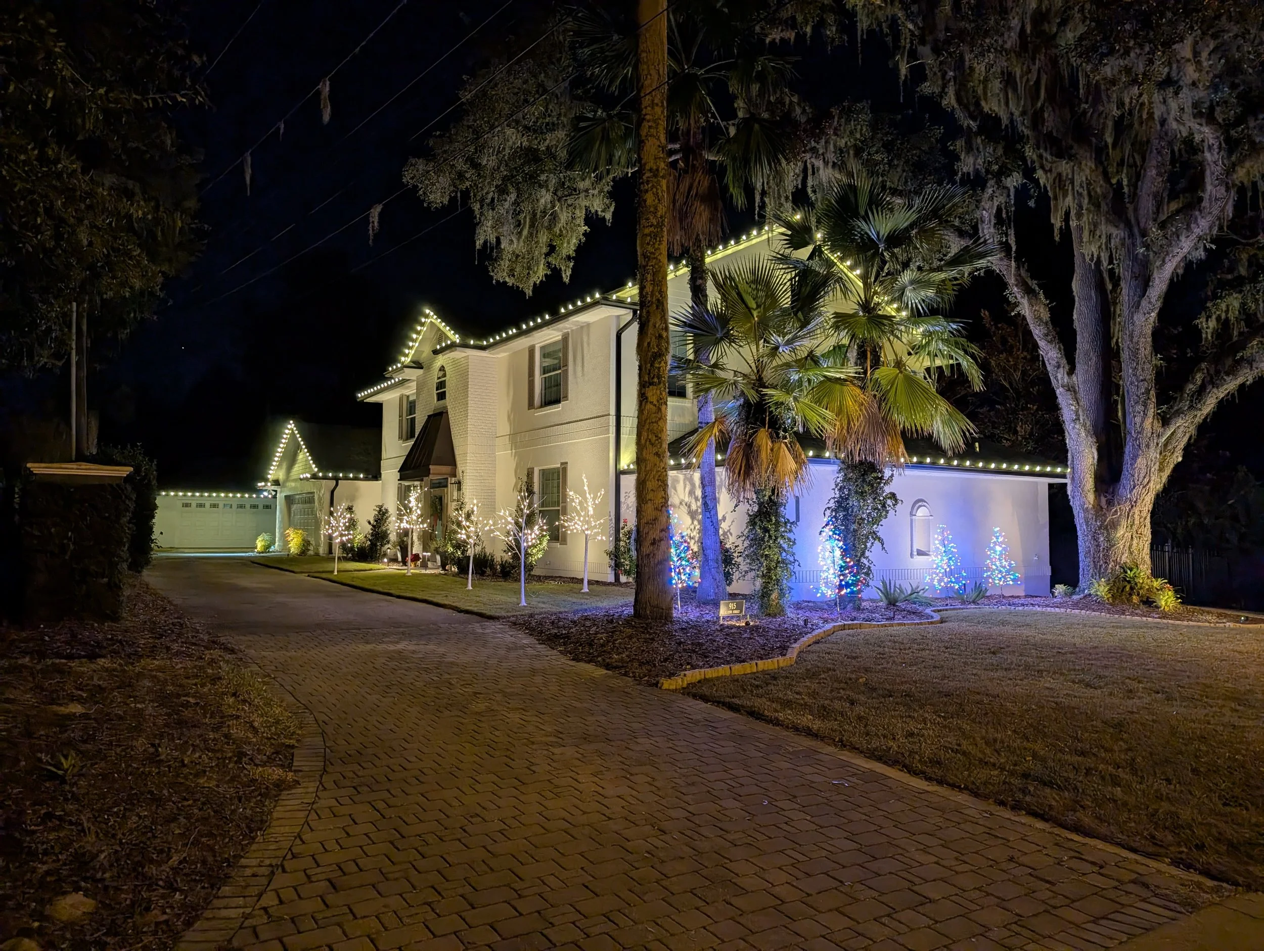 A paver driveway at night lined with palm trees lit with blue uplighting leading to a house. Orlando Christmas Lights, Winter Park Holiday Lighting, Windermere Christmas Light Installation Near Me