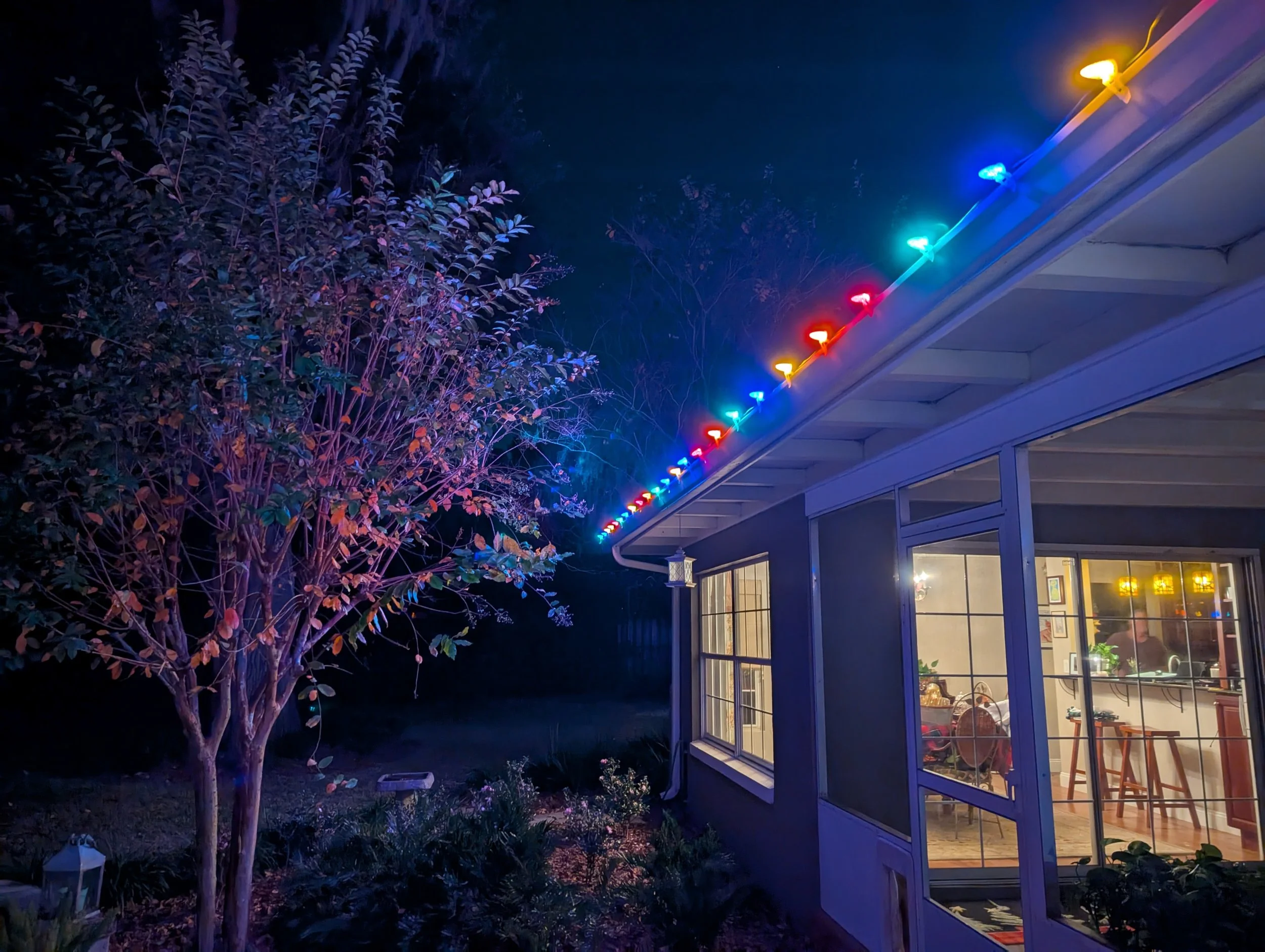 A porch roofline decorated with colorful multicolor string lights at night, with a purple-uplit tree in the foreground. Orlando Christmas Lights, Winter Park Holiday Lighting, Windermere Christmas Light Installation Near Me