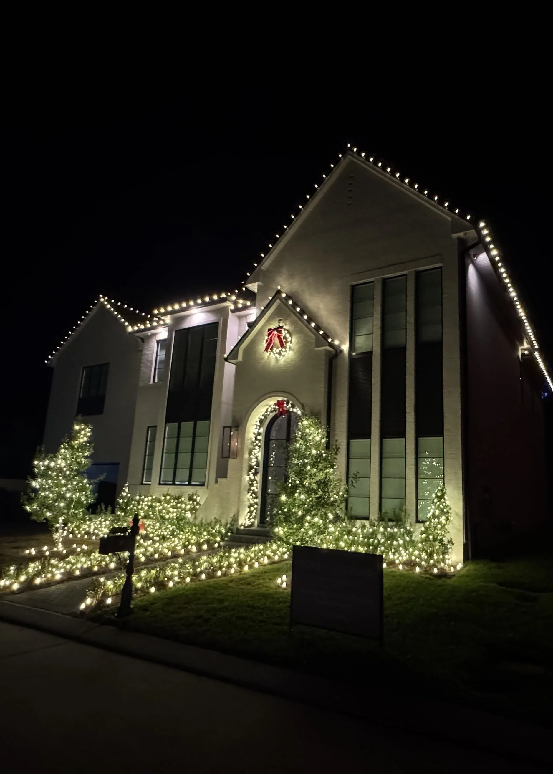 A white house at night with warm white string lights outlining the roofline, arched entryway, and wreaths on the windows. Orlando Christmas Lights, Winter Park Holiday Lighting, Windermere Christmas Light Installation Near Me