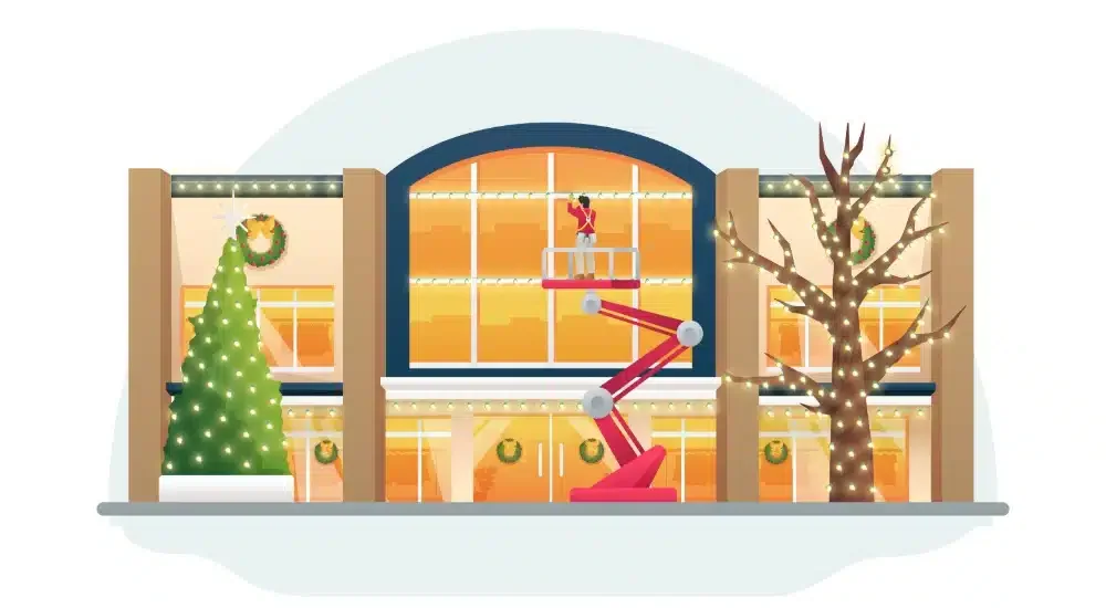 A person on a lift cleaning large windows of a decorated building with Christmas wreaths and lights during sunset.