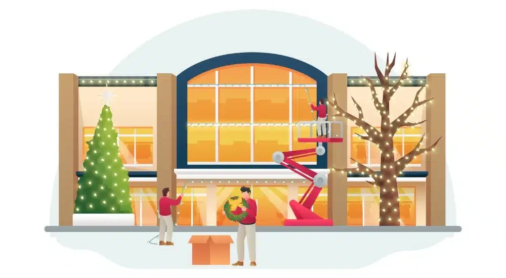 People decorating a large building with Christmas trees, lights, and holiday ornaments, with a person using a cherry picker to decorate the upper window.