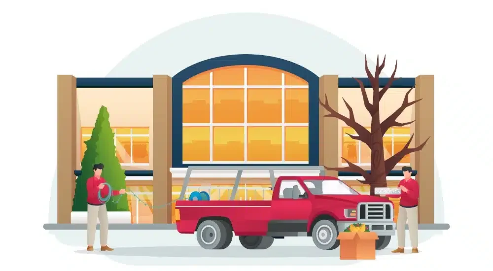 People unloading supplies from a red pickup truck in front of a large building with big windows and an arched entrance, with a leafless tree and a tall green bush nearby.