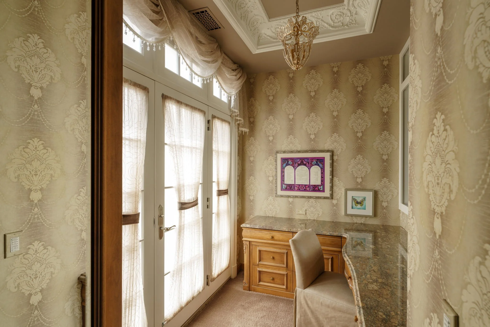 Interior view of a living space with ornate wallpaper, large windows with sheer curtains, a chandelier hanging from the ceiling, framed artwork on the wall, a granite countertop, and a wooden cabinet.
