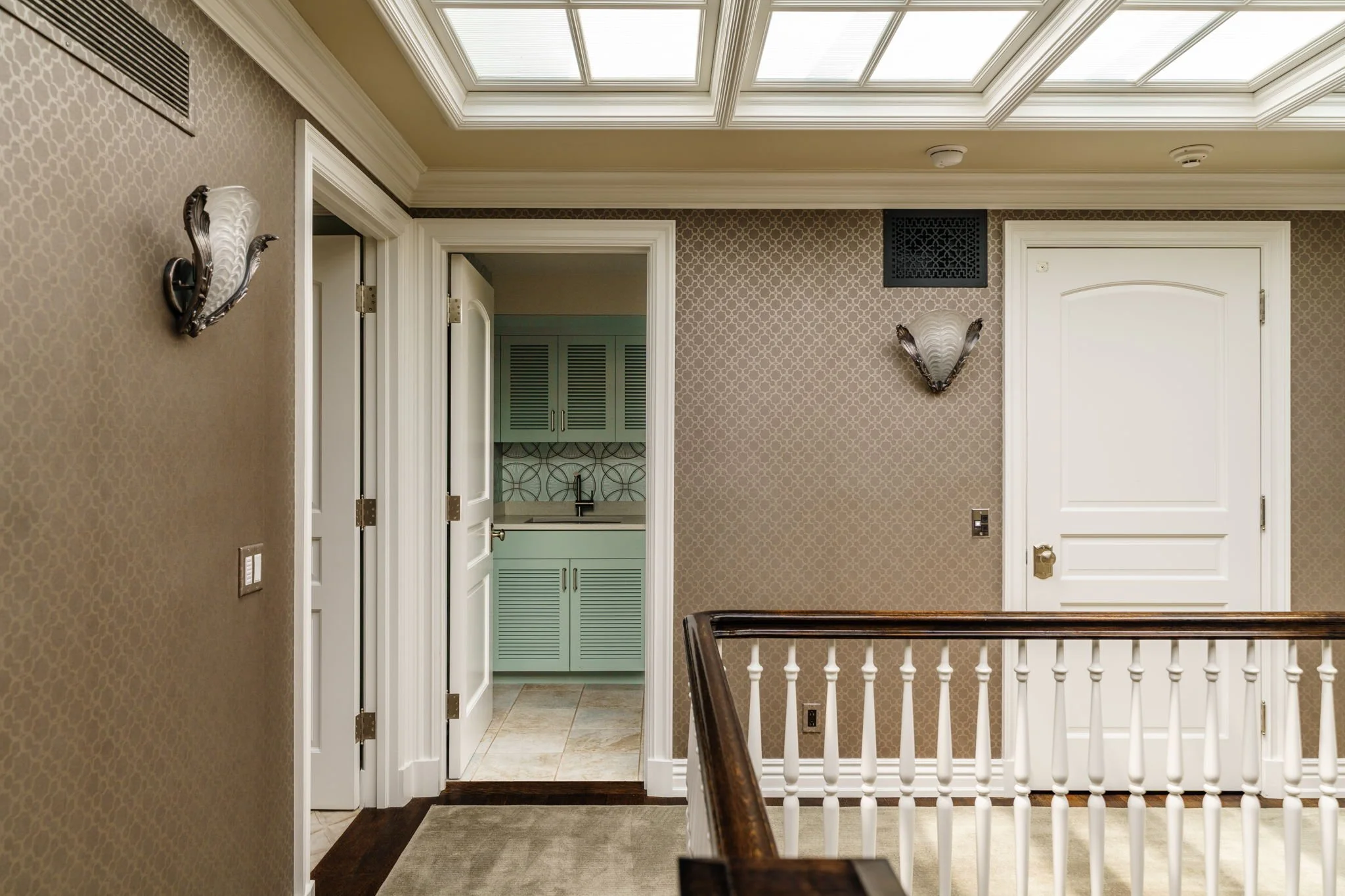 An interior hallway with beige patterned wallpaper, a white door, and a small kitchenette with light green cabinets visible through an open door. There's a staircase with a dark wooden handrail in the foreground, ceiling lights, and a skylight.