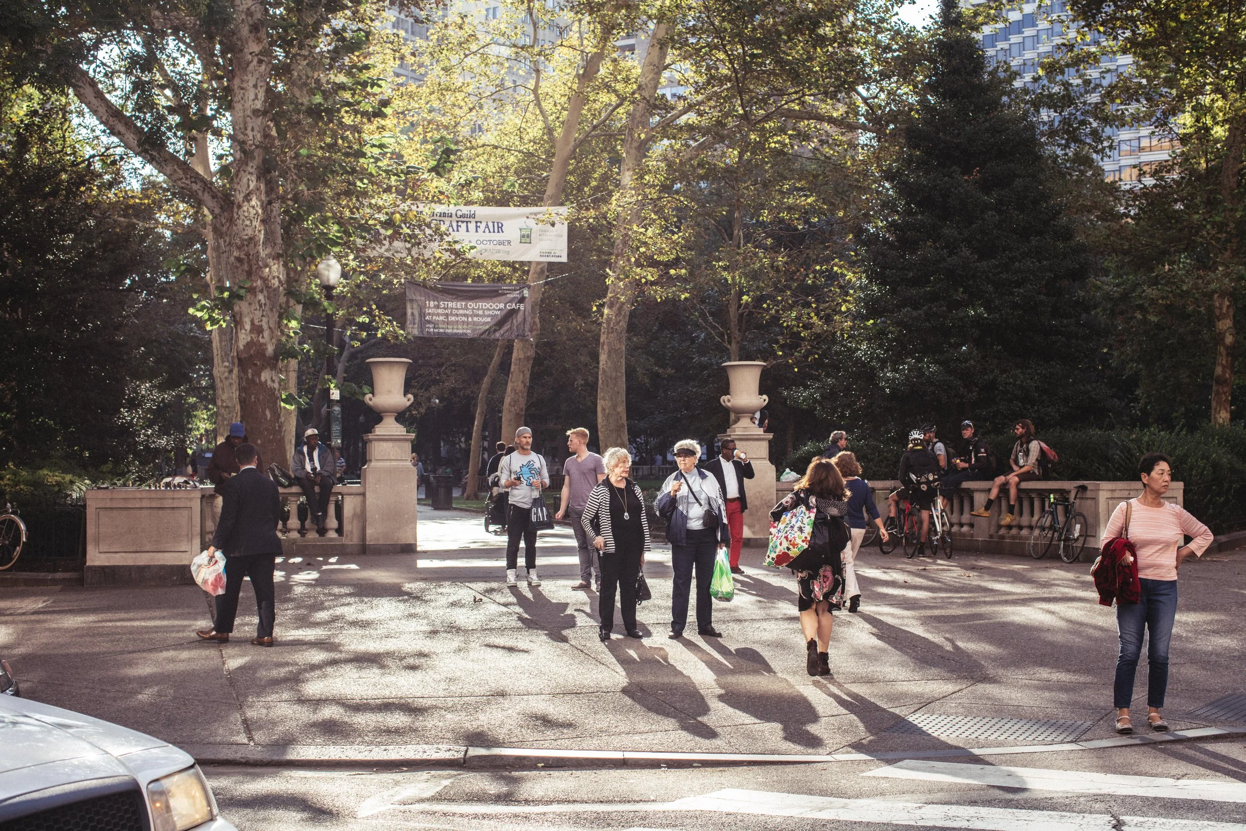 A busy outdoor urban park scene with people walking, sitting, and standing under trees. There are bicycles, a car partially visible in the foreground, and banners hanging between trees advertising a craft fair and outdoor cafe. The background features tall buildings.