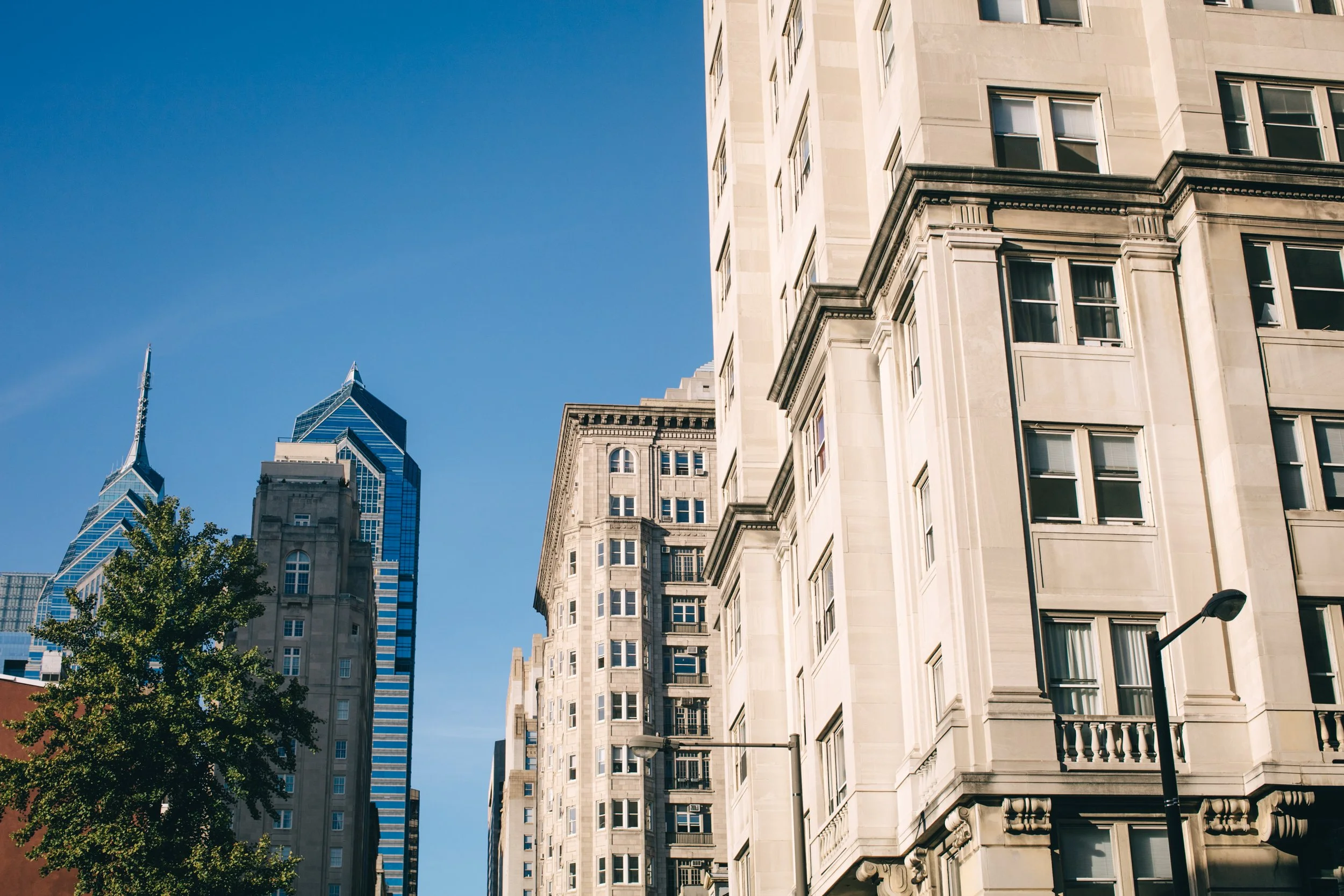 Street view of tall buildings in a city, with a bright blue sky and a tree in the foreground.