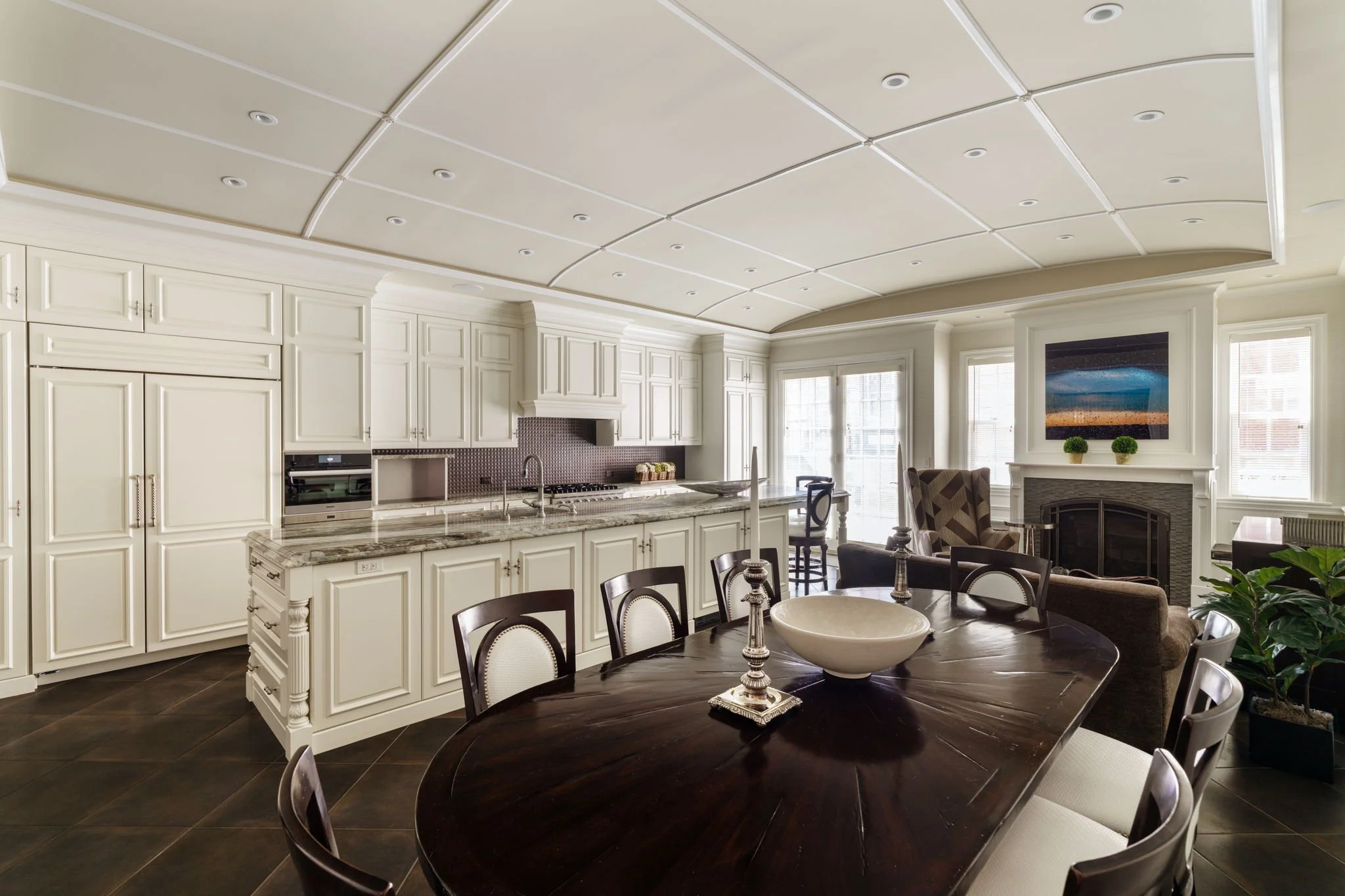 A spacious kitchen with white cabinets and a large dark wooden dining table with chairs. The kitchen features a marble countertop, built-in appliances, and a tiled backsplash. There is a fireplace with two small potted plants above it in the adjacent