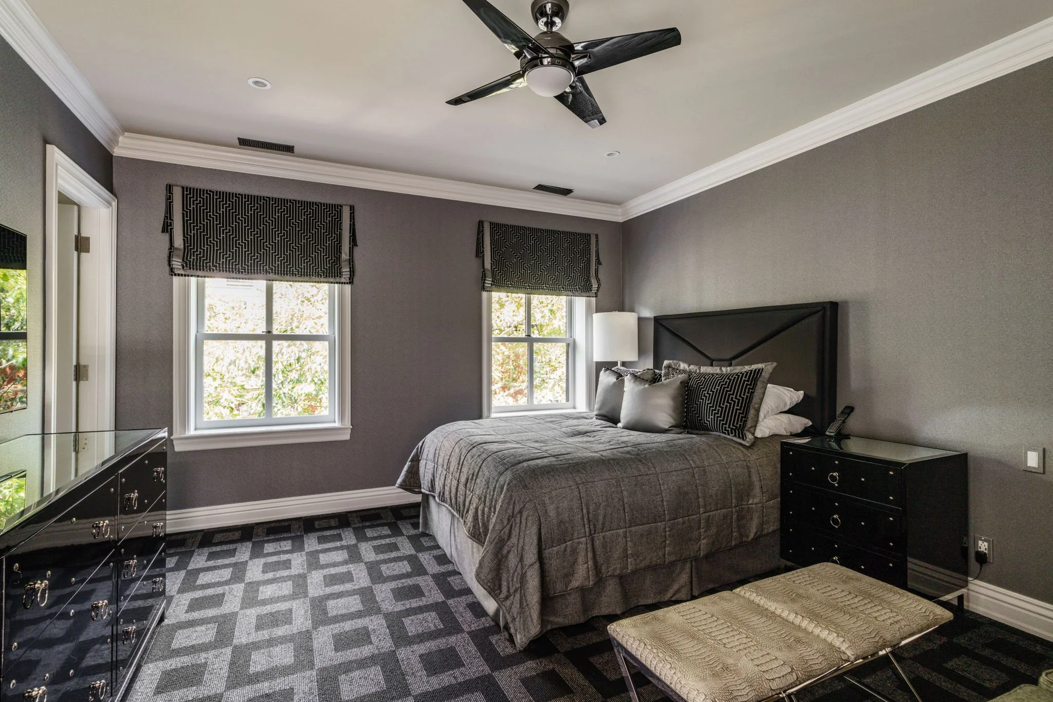 A bedroom with a bed, two nightstands, and two windows with patterned black and white curtains. The room has crown molding and a ceiling fan. The walls are painted gray, and there is a patterned carpet on the floor.