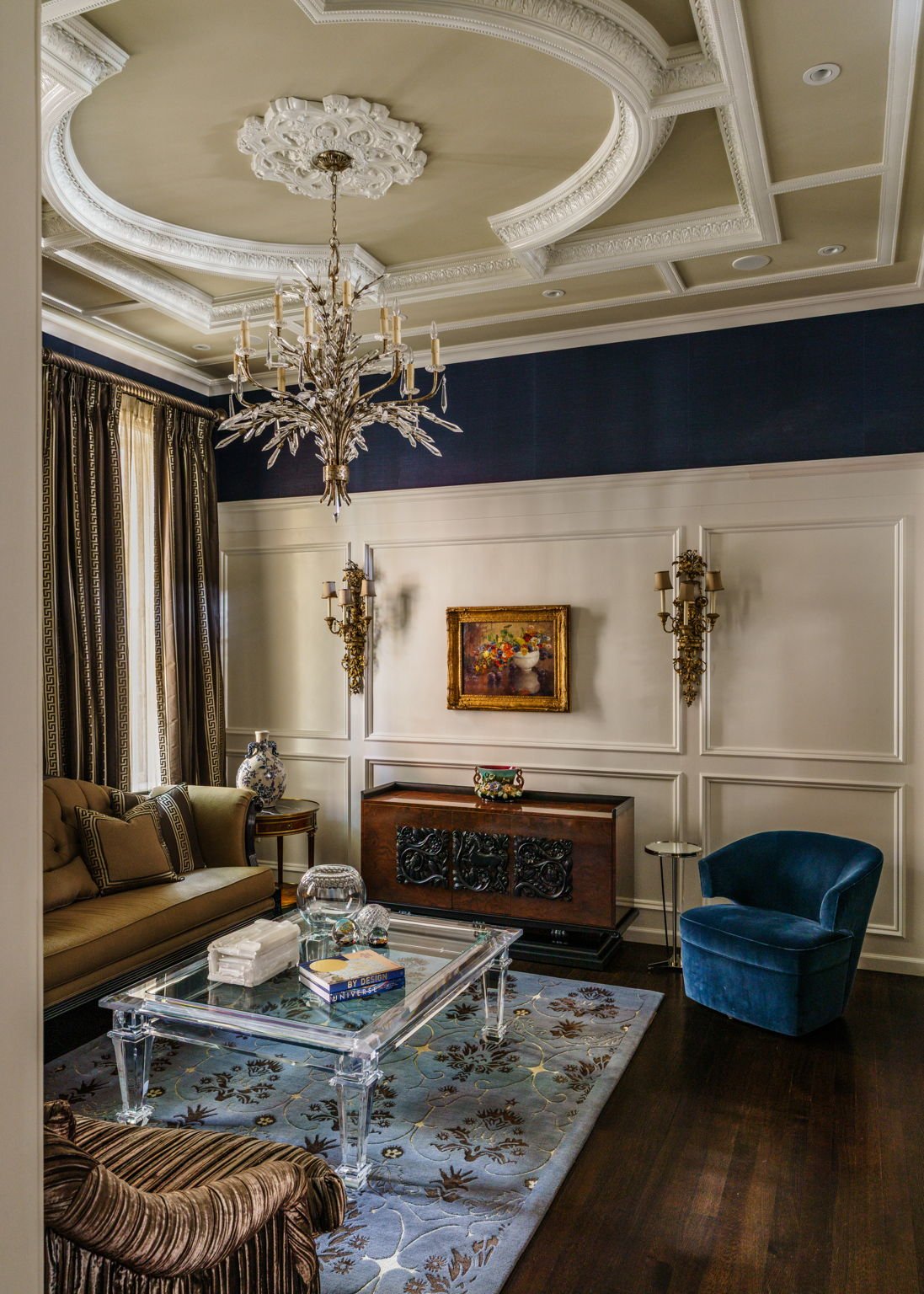 Elegant living room with ornate ceiling, crystal chandelier, wall sconces, framed painting, vintage furniture, glass coffee table, and decorative rug.