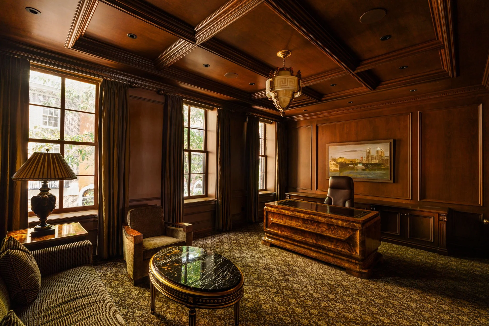 Wood-paneled office with large windows, curtains, a chandelier, leather chairs, and brass-trimmed furniture.