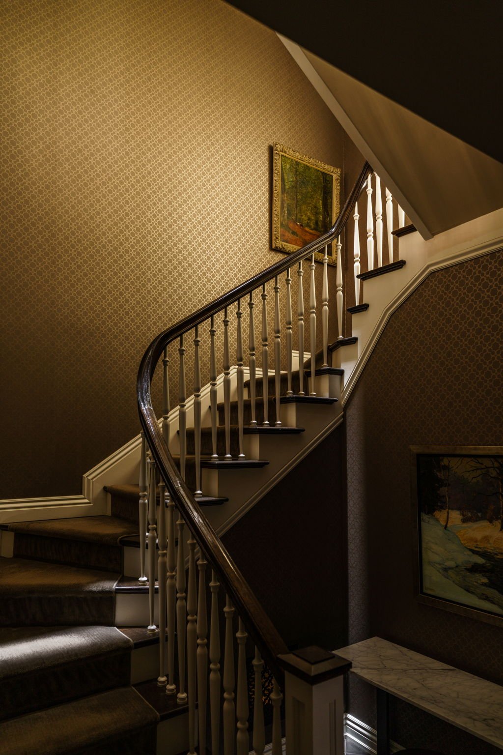 Interior view of a staircase with dark wooden handrail and white balusters, wallpapered walls, framed landscape paintings, and a marble-topped table.