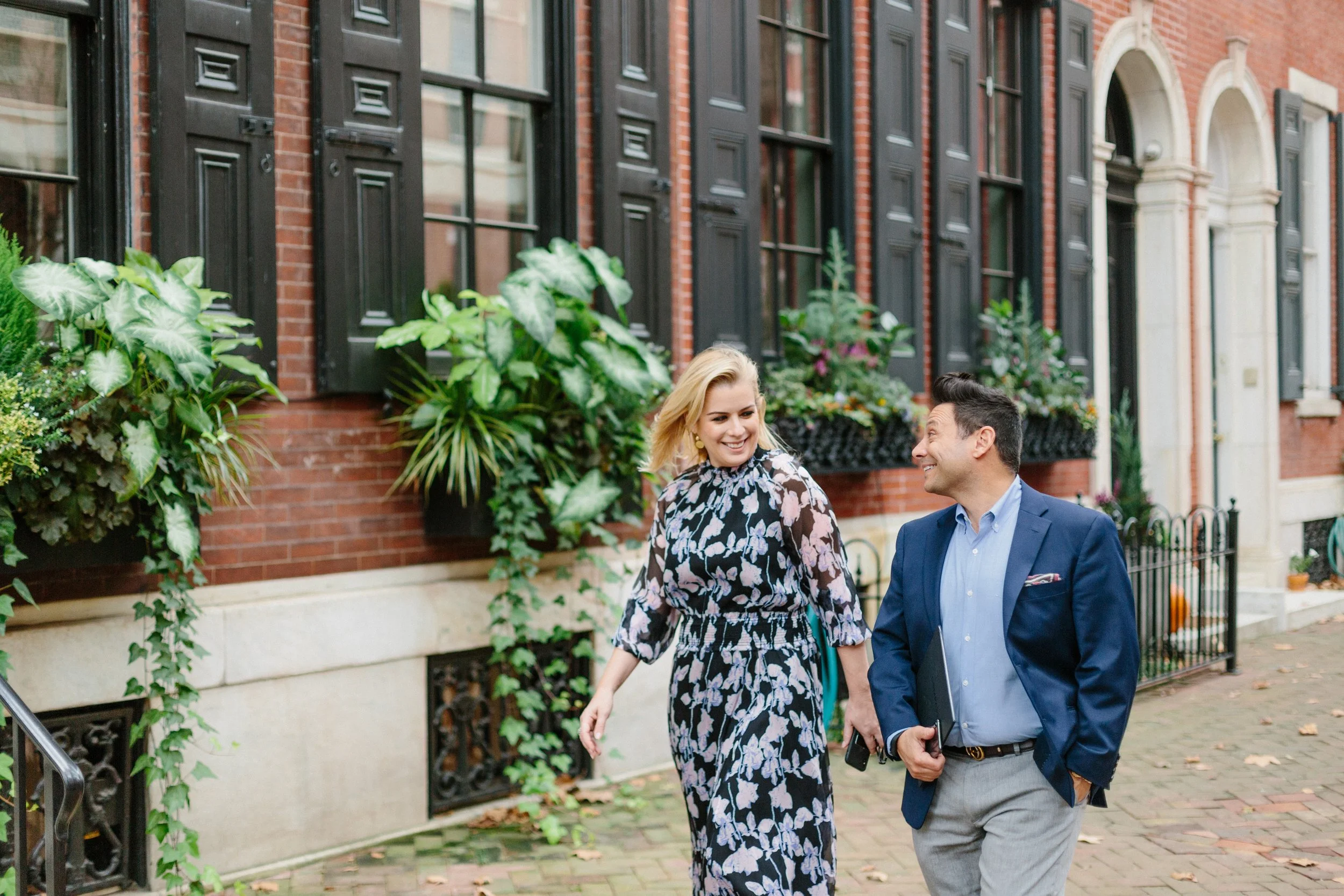Two people, a woman in a floral dress and a man in a blue blazer and gray pants, walking down a city sidewalk in front of a row of brick townhouses with black shutters and lush green plants in window boxes, smiling and engaging in conversation.