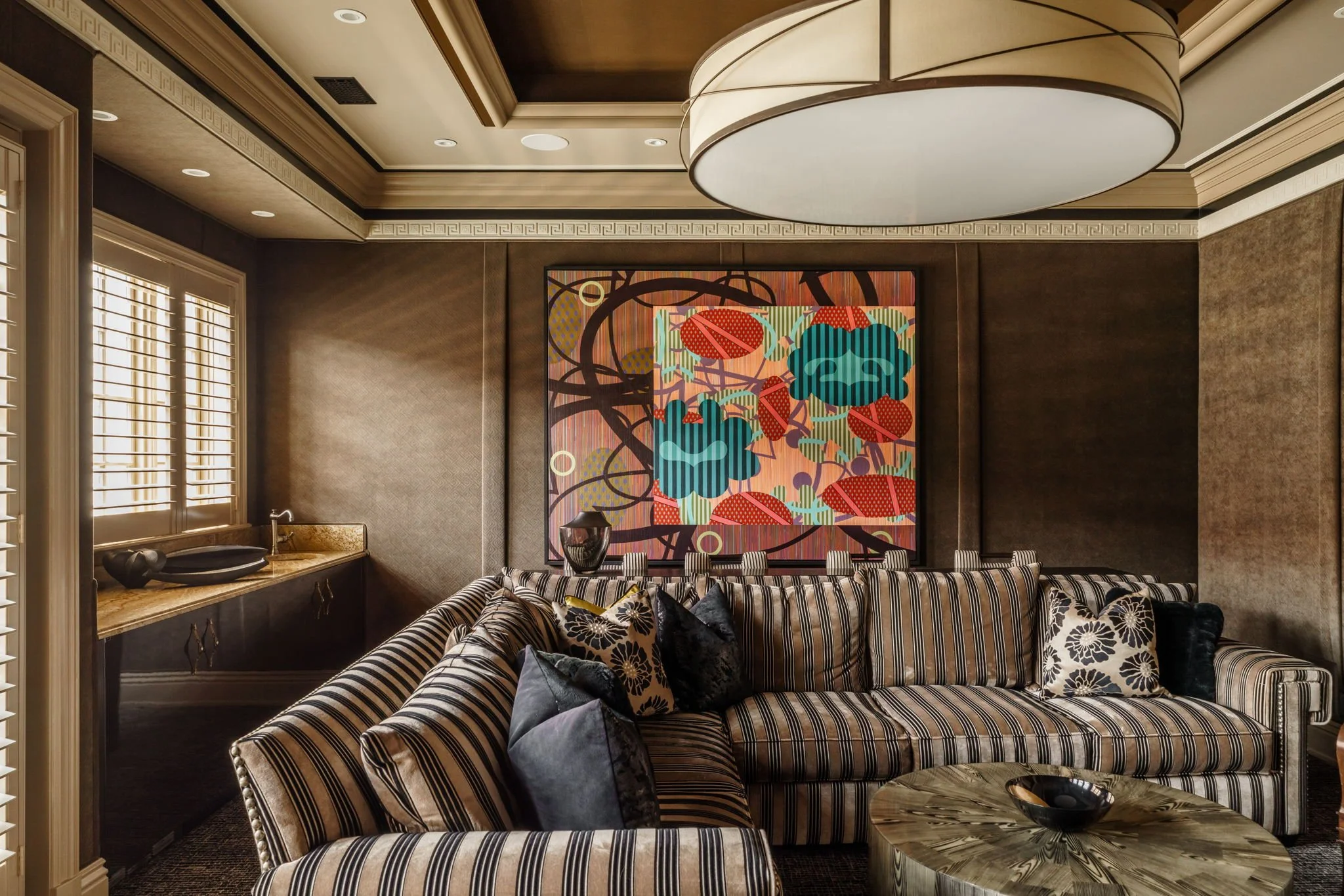 An elegant living room with a striped sofa, black and white pillows, and a large colorful abstract painting on the wall. There's a marble-top side table with a black bowl and a vase, and window shutters on the left letting in natural light.