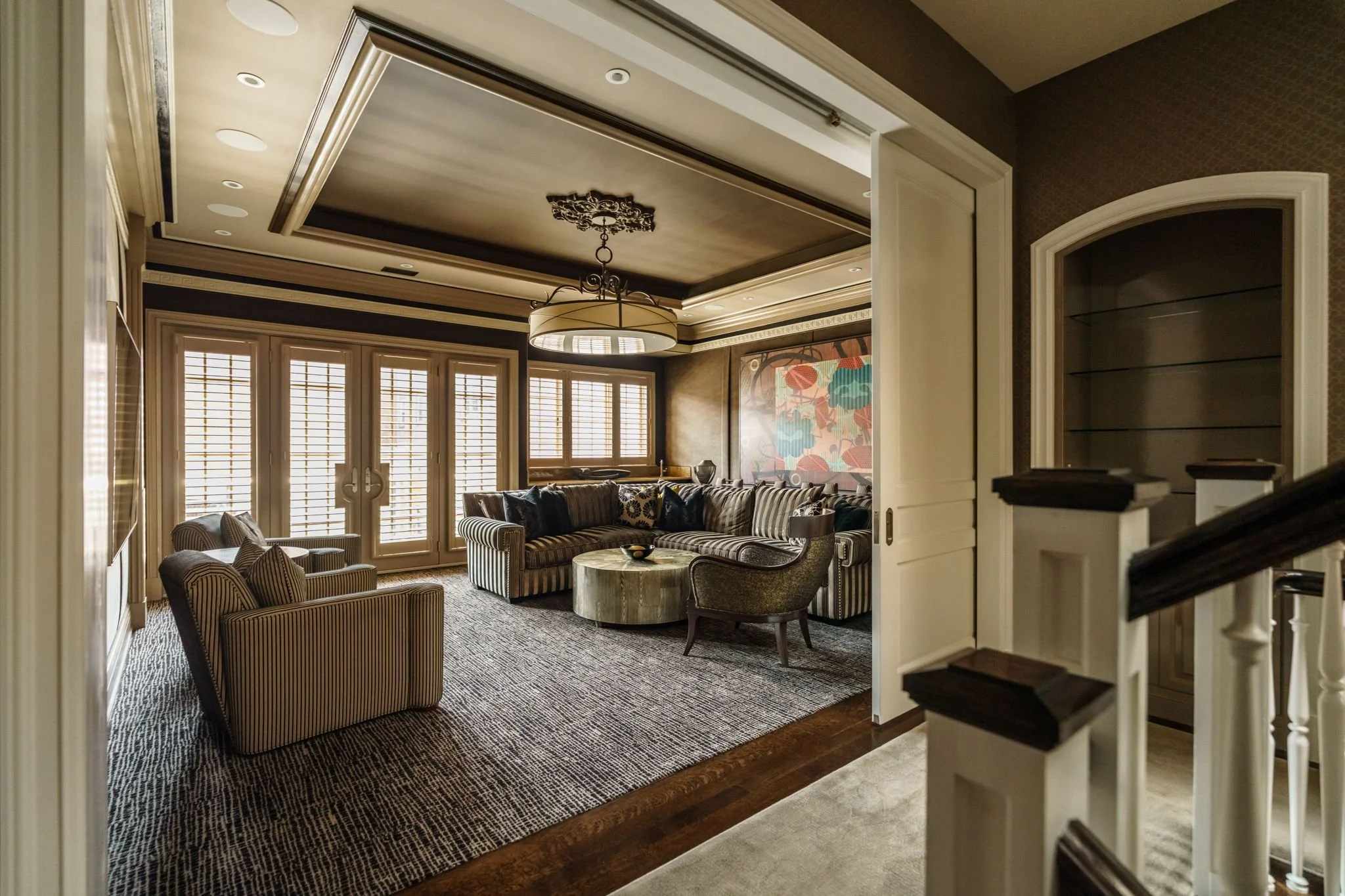 Elegant living room with striped sofas, armchairs, a round coffee table, large windows with shutters, decorative wallpaper, and artwork on the wall. A chandelier hangs from the ceiling.