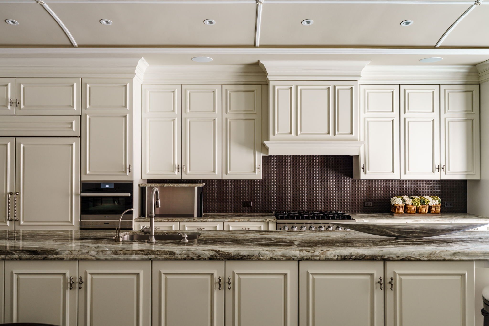 A kitchen with cream-colored cabinets, a granite countertop, and a dark patterned backsplash. A built-in oven is visible, along with a stainless steel sink and faucet. There are small decorative baskets of flowers on the counter.