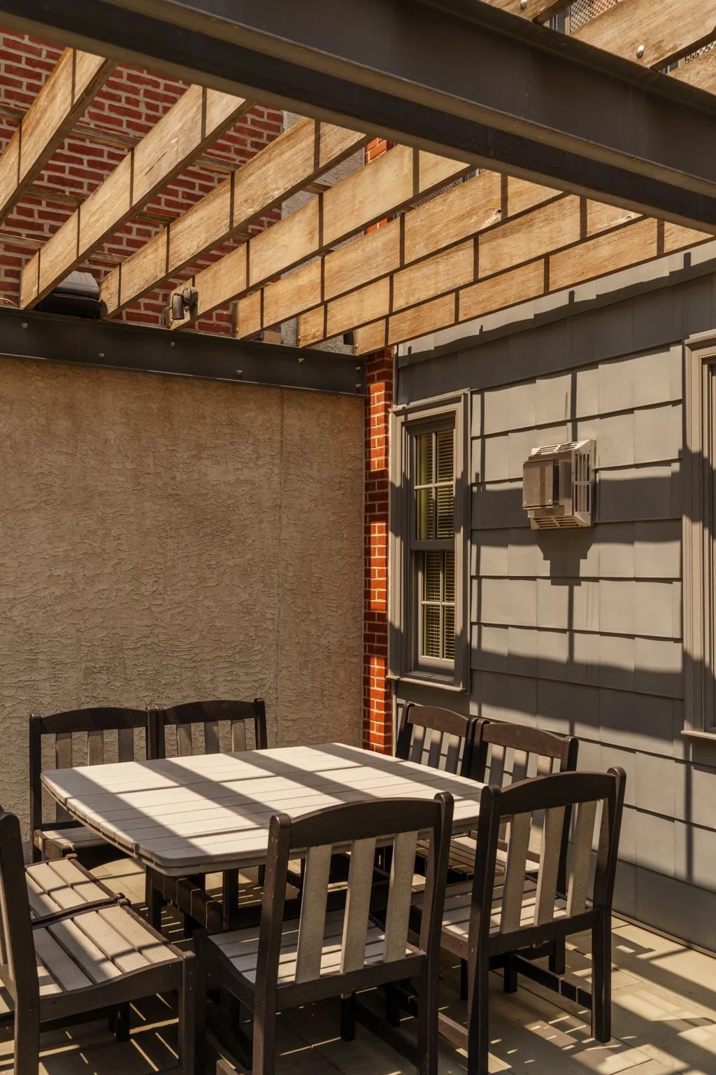 An outdoor patio area with a white table and six black chairs, surrounded by walls with one having a window and an air conditioning unit, and a partially built wooden pergola overhead with sunlight casting shadows.