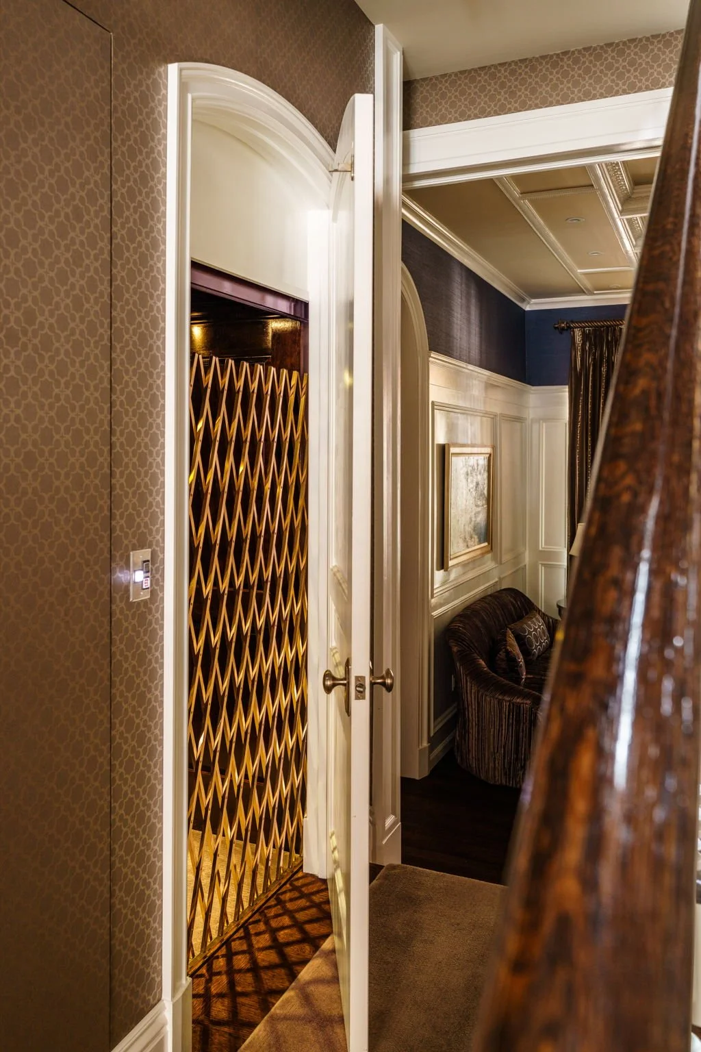 View into a luxurious hotel suite with a partially open door revealing a gold-colored foldable security gate, elegant wall paneling, dark blue & beige walls, a framed painting, a plush armchair with decorative pillows, and patterned curtains.