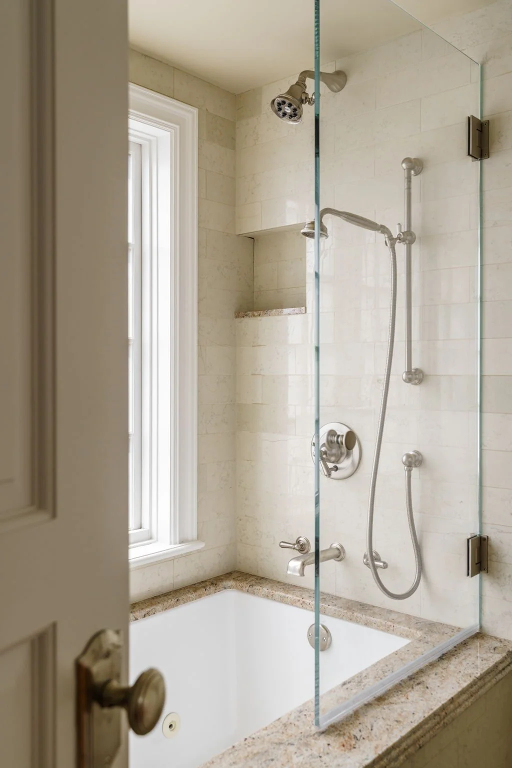 A bathroom shower with a glass partition, a showerhead, a handheld shower, a built-in shelf, and a window with a white frame.