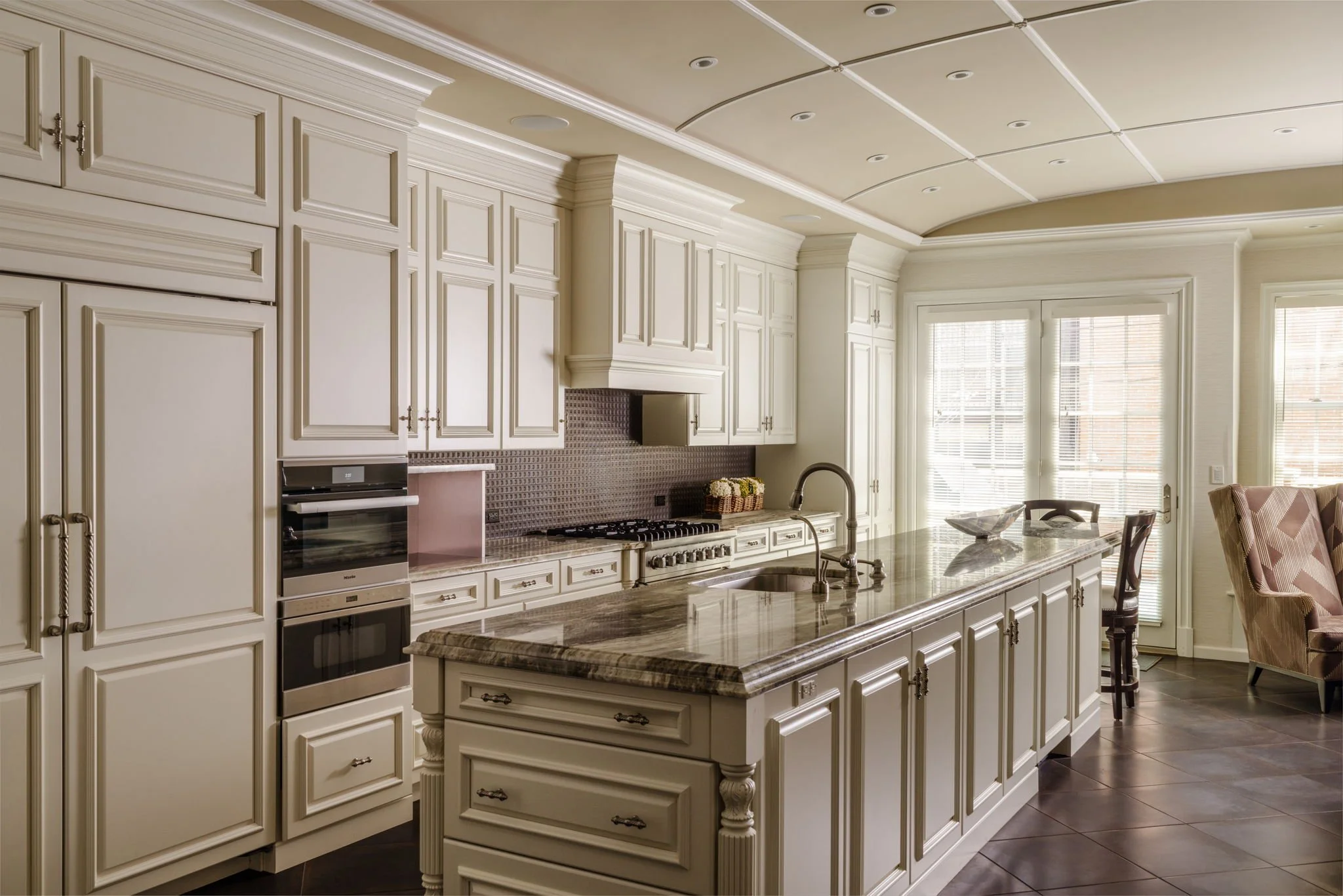 Cream-colored kitchen with cabinetry, a granite island, a stove and oven, a window with blinds, and a sitting area with a chair.