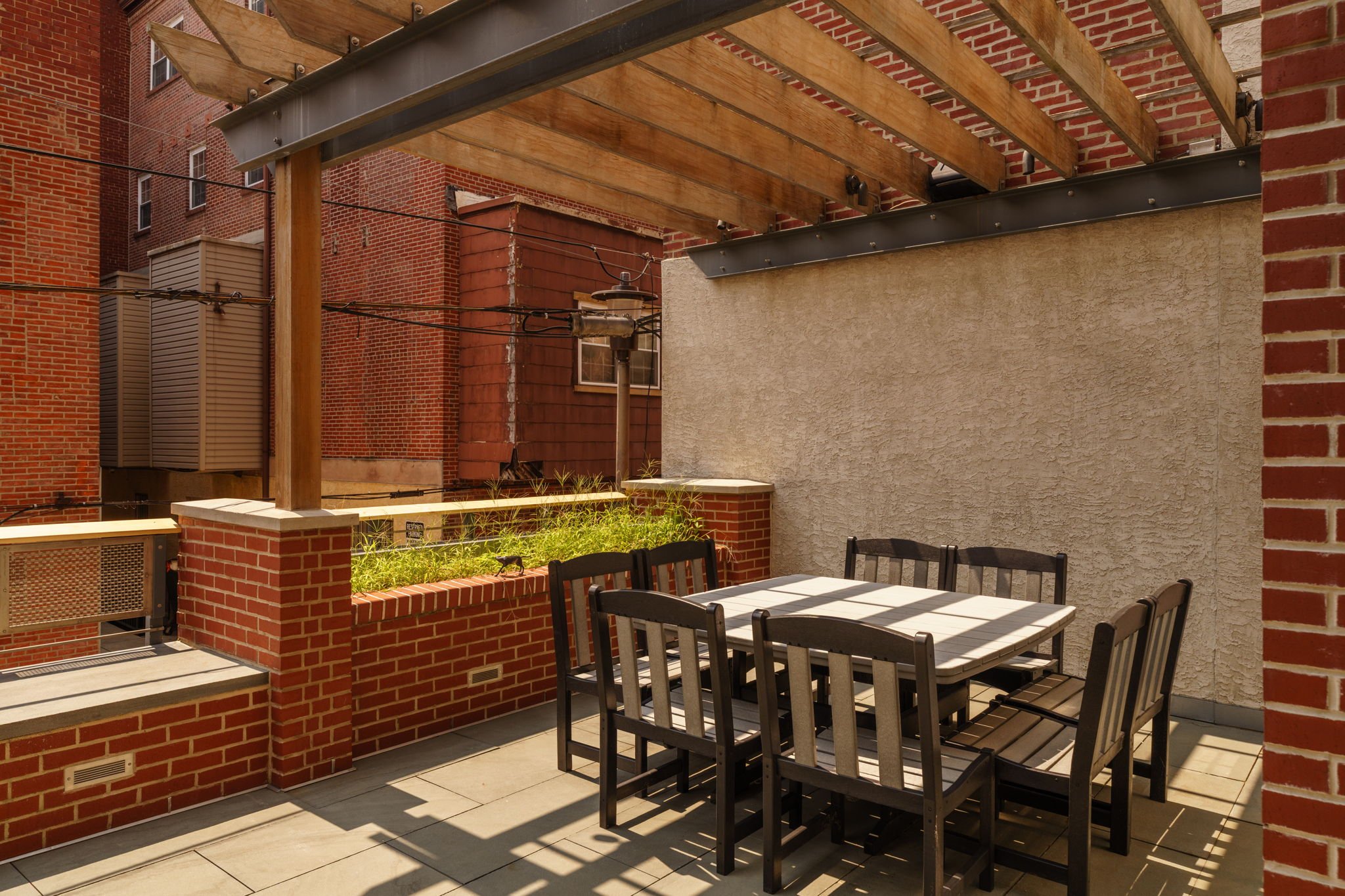 A rooftop patio area with a white table and six black chairs, surrounded by brick walls and buildings. There is a small raised garden bed with green plants, and a string of black wires or cables overhead. Some chairs are partially in the shadow.