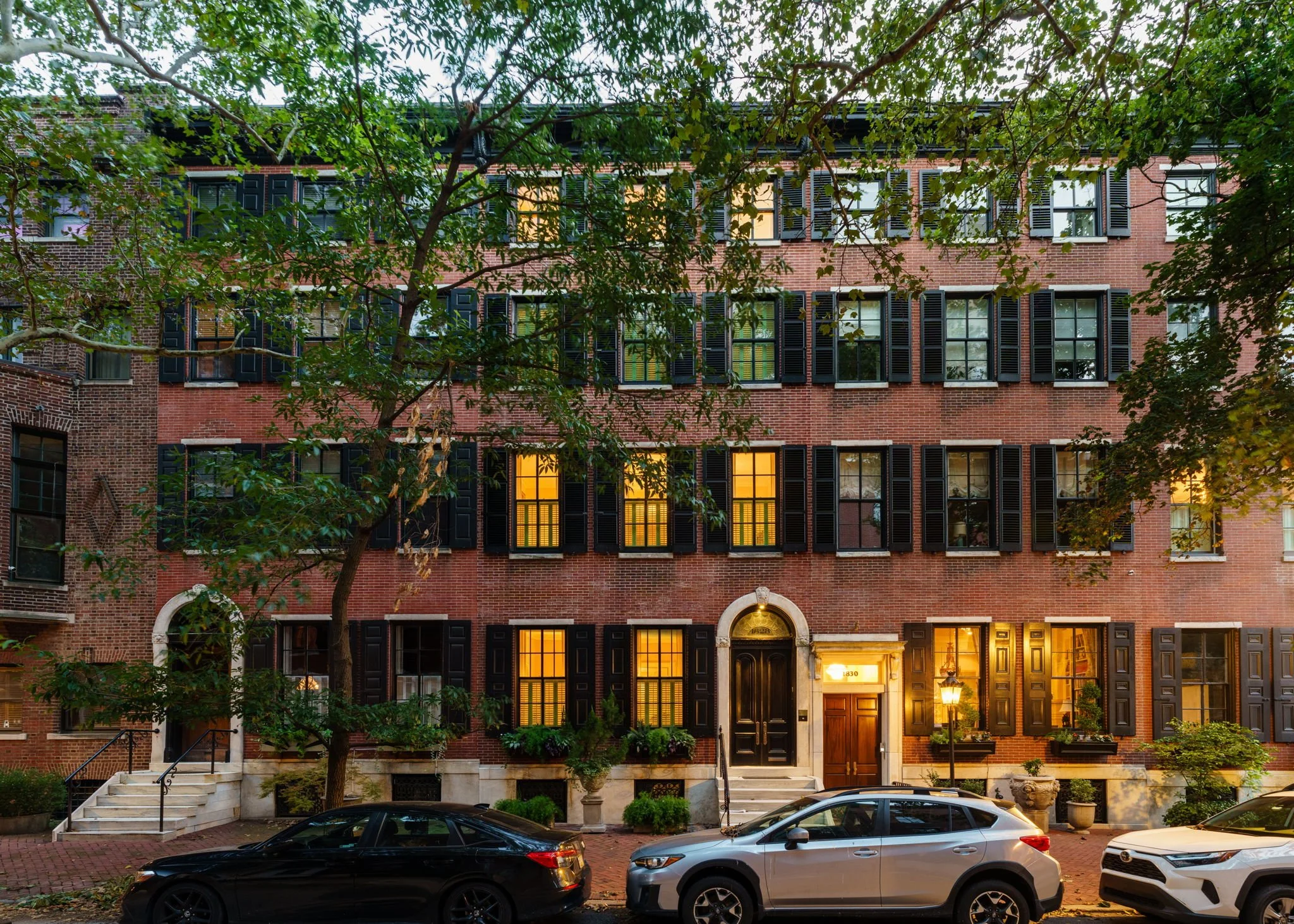 A brick apartment building with multiple windows and black shutters, some illuminated from inside. The entrance has a black door with a gold handle and a lit lamp nearby. There are several cars parked in front, and trees partially cover the building.
