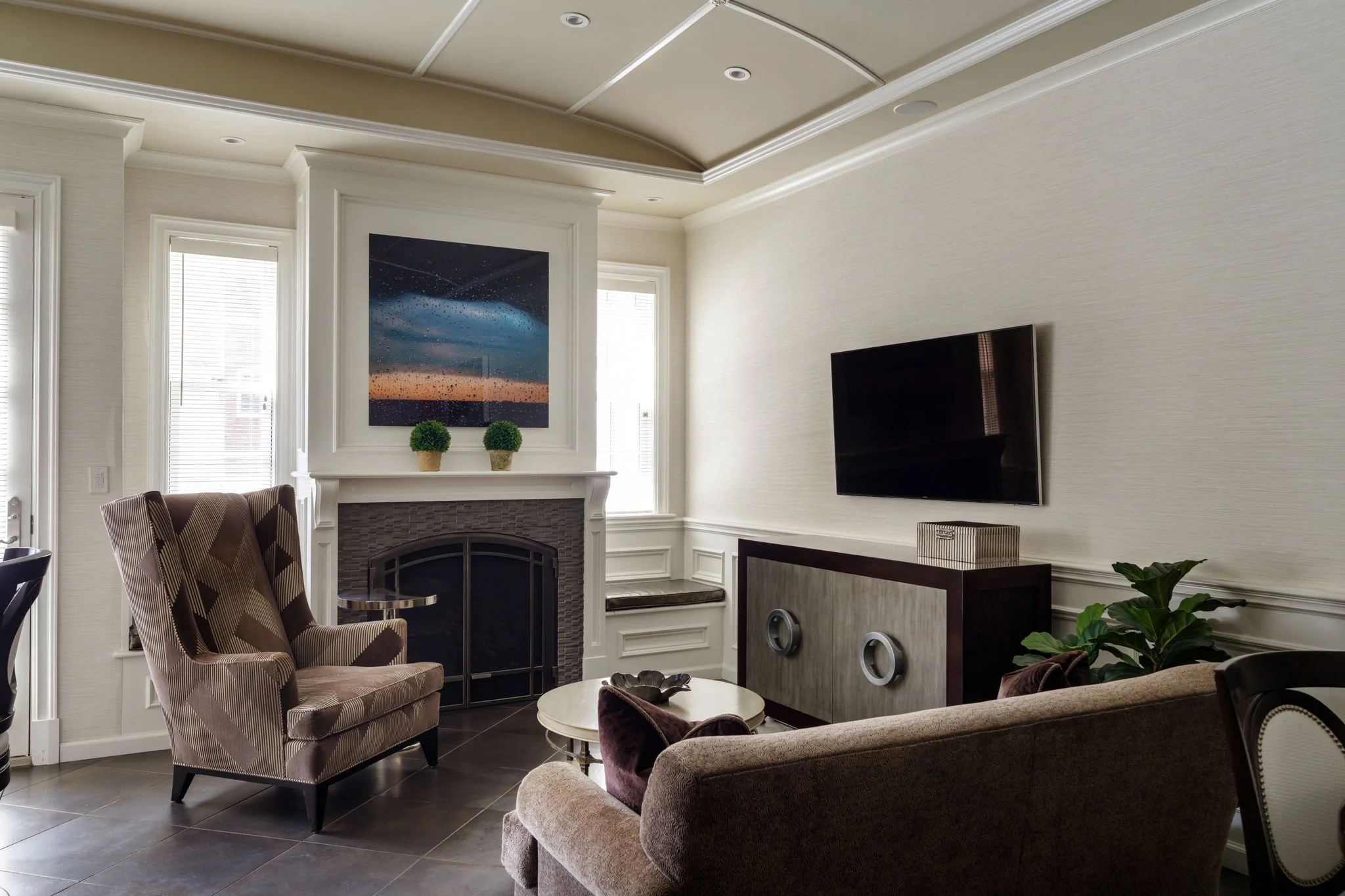 A living room with a fireplace, a large flat-screen TV on the wall, and two windows with white blinds. There is a patterned armchair, a beige sofa, a small round coffee table, and some decorative items including two potted plants and wall art depicti