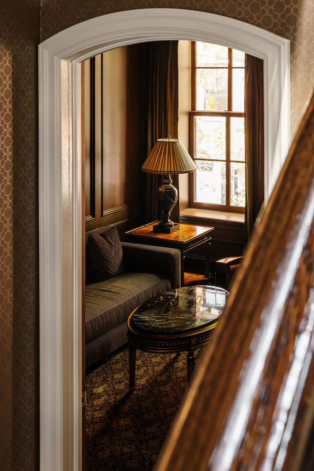 A cozy, elegant room with a window allowing natural light, dark wood paneling, a striped armchair, a black marble-topped coffee table, a table lamp, and patterned carpet.