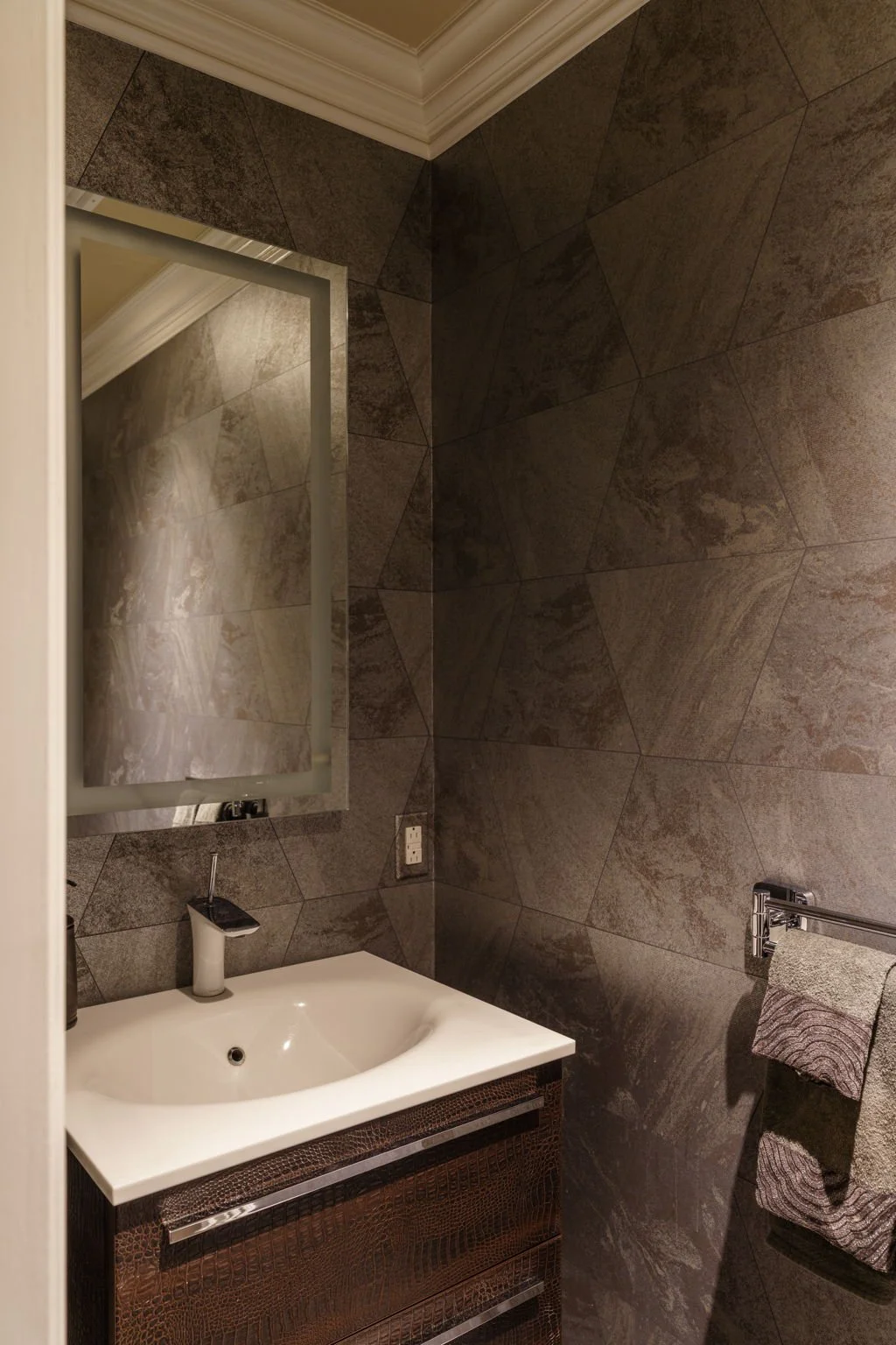 A modern bathroom with dark gray textured tile walls, a white sink with a cabinet beneath, a rectangular mirror, a towel rack with two patterned towels, and an electrical outlet.