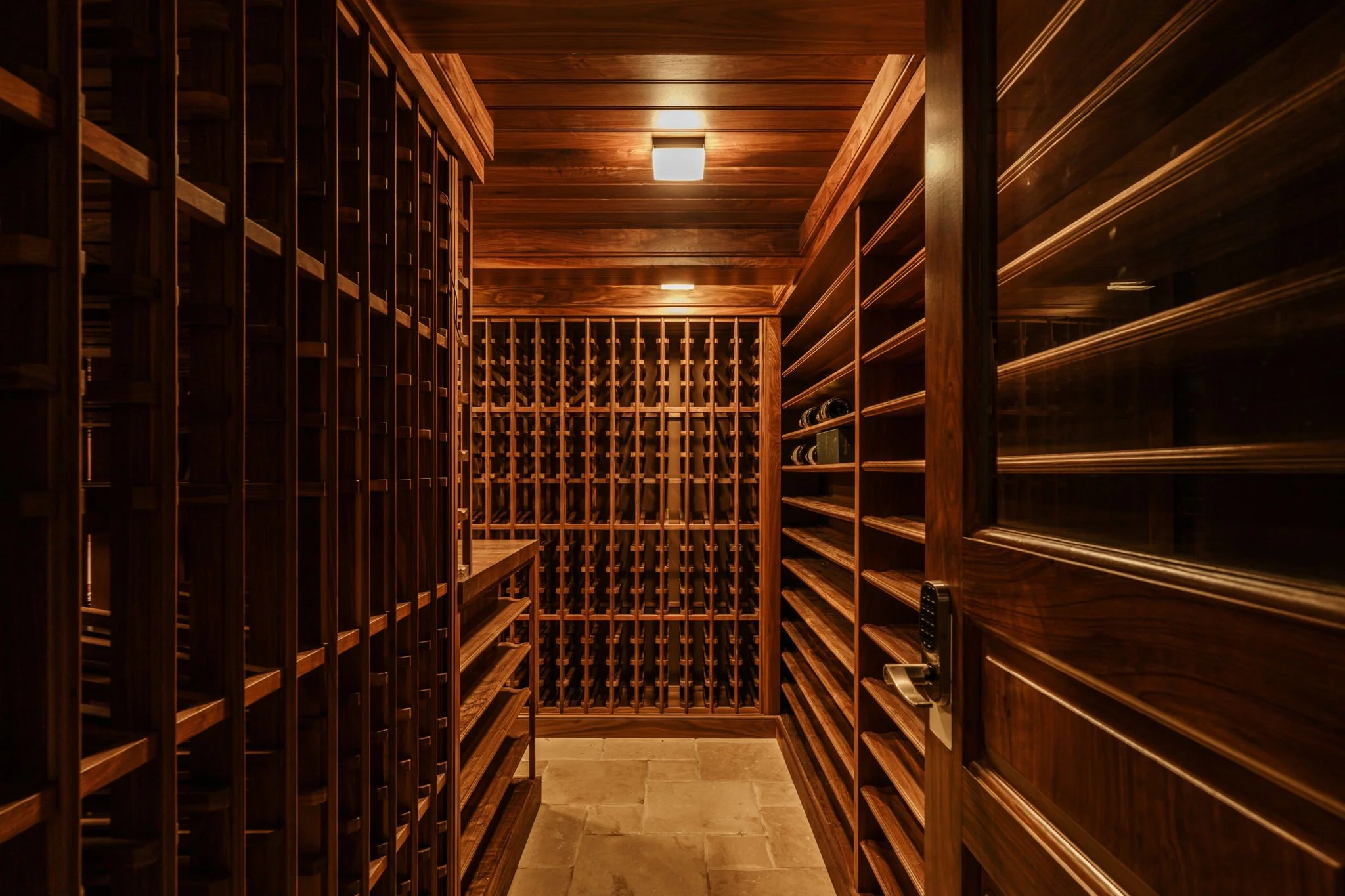 A wooden wine cellar with shelves on both sides and a wine rack at the back, illuminated by ceiling lights.