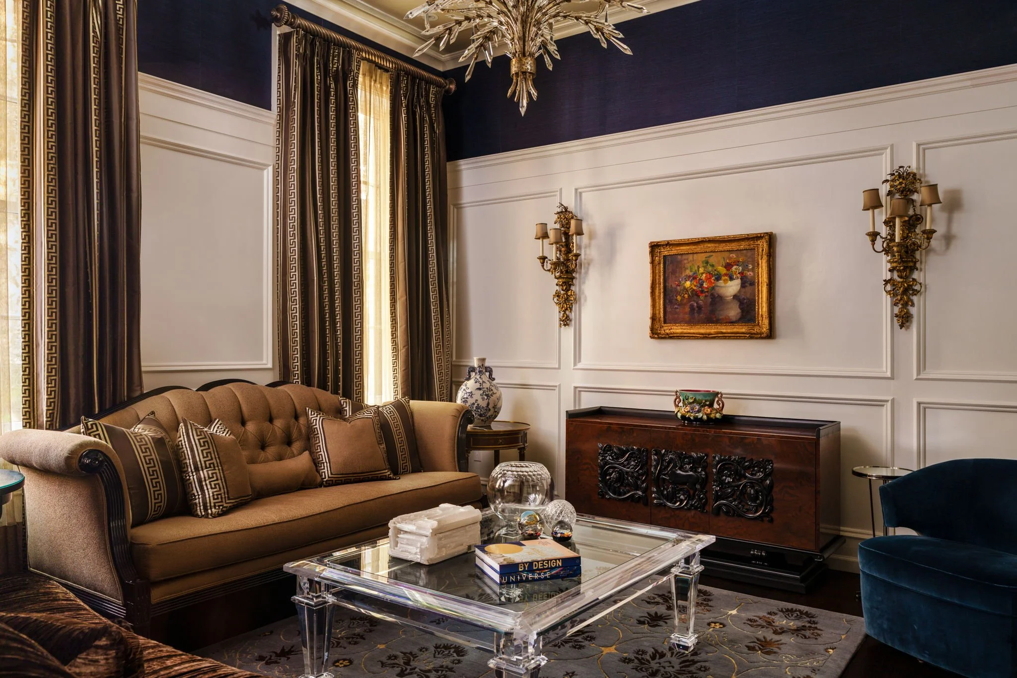 A luxurious living room with navy blue walls, white decorative wall panels, and elegant gold wall sconces. There are large windows with dark, patterned drapes, and a chandelier hanging from the ceiling. The room features a beige sofa with matching cu