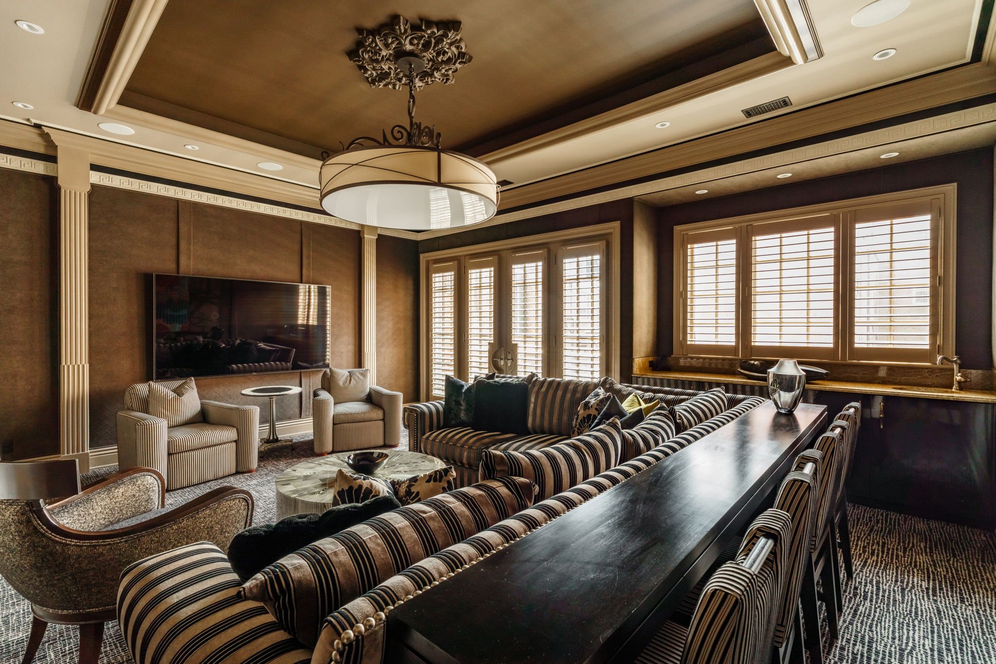 A ceiling with ornate molding and a large round chandelier. Multiple windows with white blinds allow natural light into a living room with striped and solid upholstered furniture, a wall-mounted flat-screen TV, and a dark dining table with striped ch