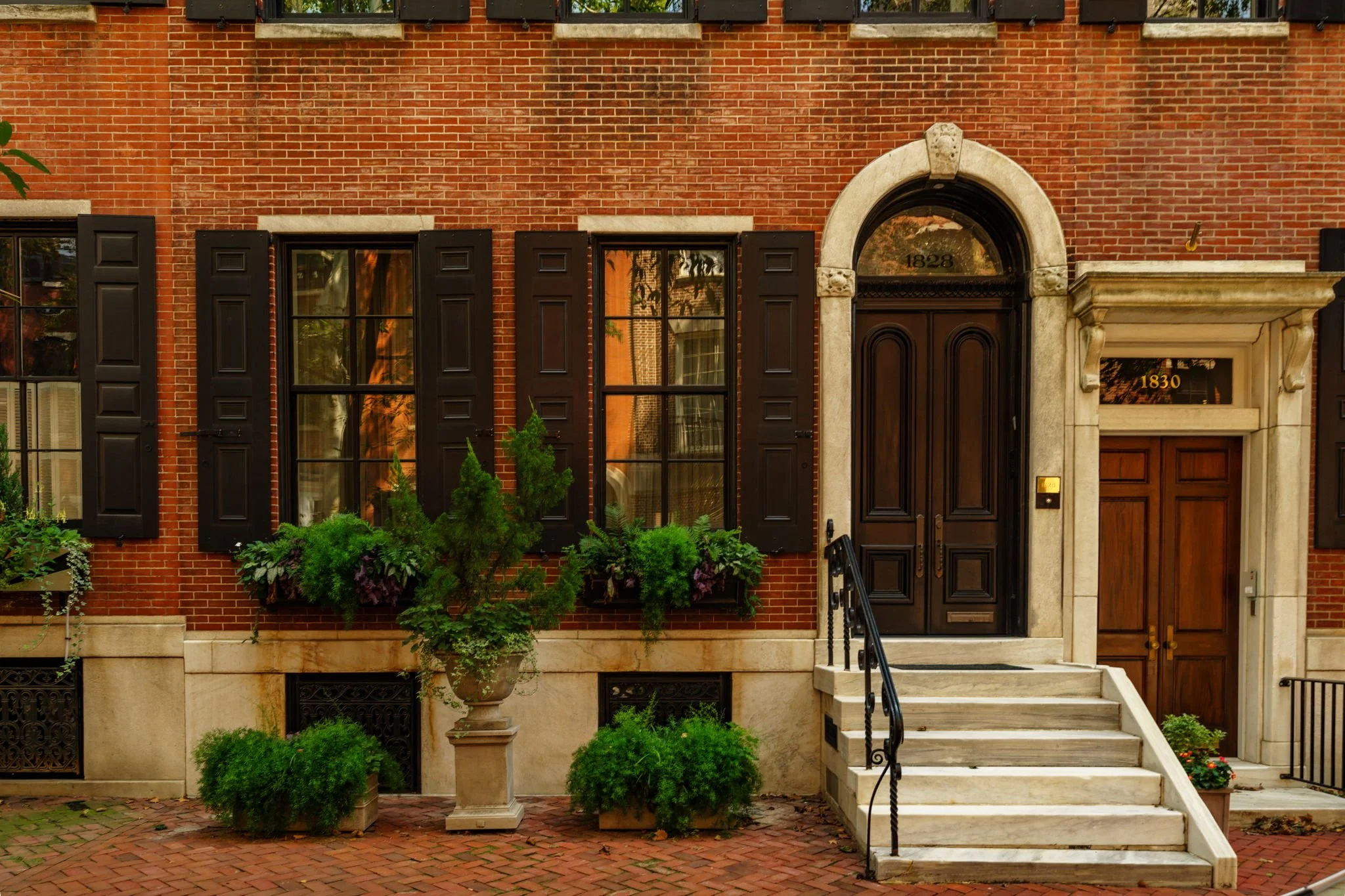 Brick townhouse with black window shutters, arched doorway with the number 1828, stone trim, a staircase with wrought iron railing, and green bushes and plants in front.
