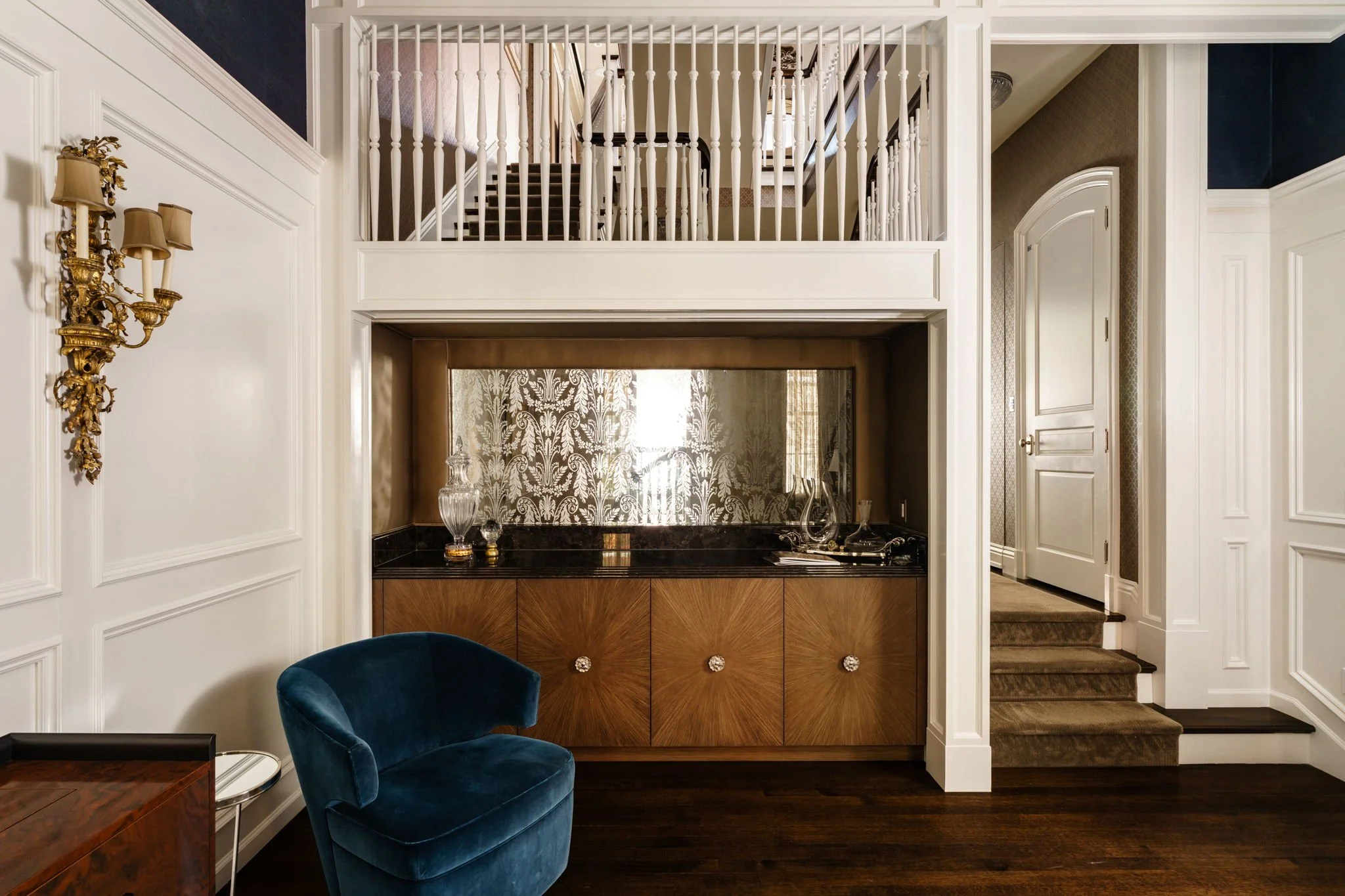 Interior view of a living room with elegant decor, including a blue velvet chair, a wooden cabinet with decorative handles, a gold wall sconce, and a staircase with white banisters leading to an upper floor.